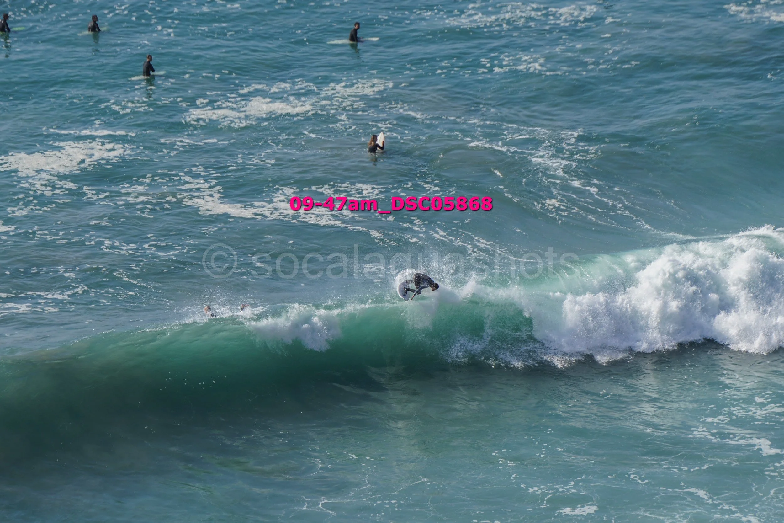 A person in a wetsuit riding a surfboard on a breaking wave while several other surfers are visible in the ocean in the background.