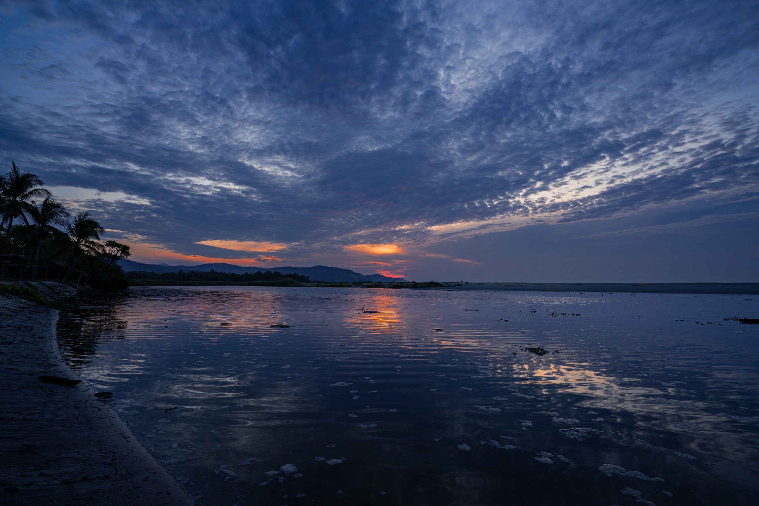 A scenic view of a sunset over a body of water, with a partly cloudy sky and silhouettes of palm trees on the left side.