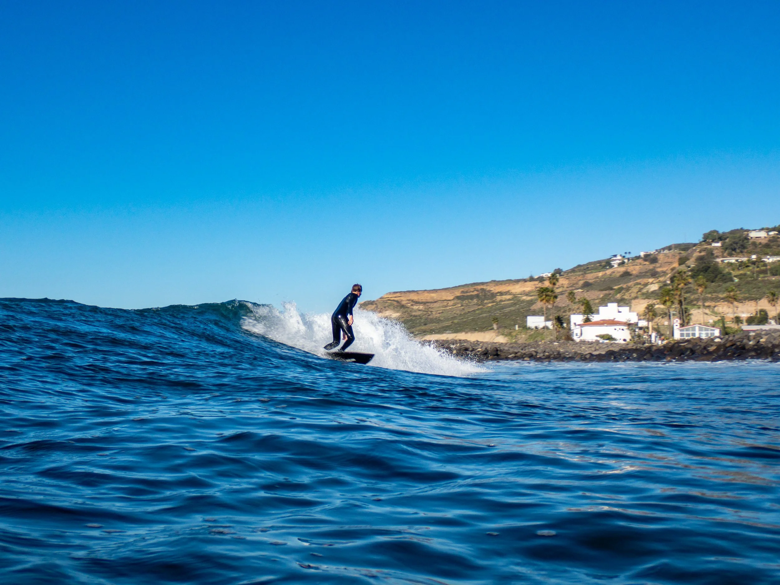 A person in a wetsuit riding a surfboard on a wave near the shoreline with houses and hilly terrain in the background under a clear blue sky.