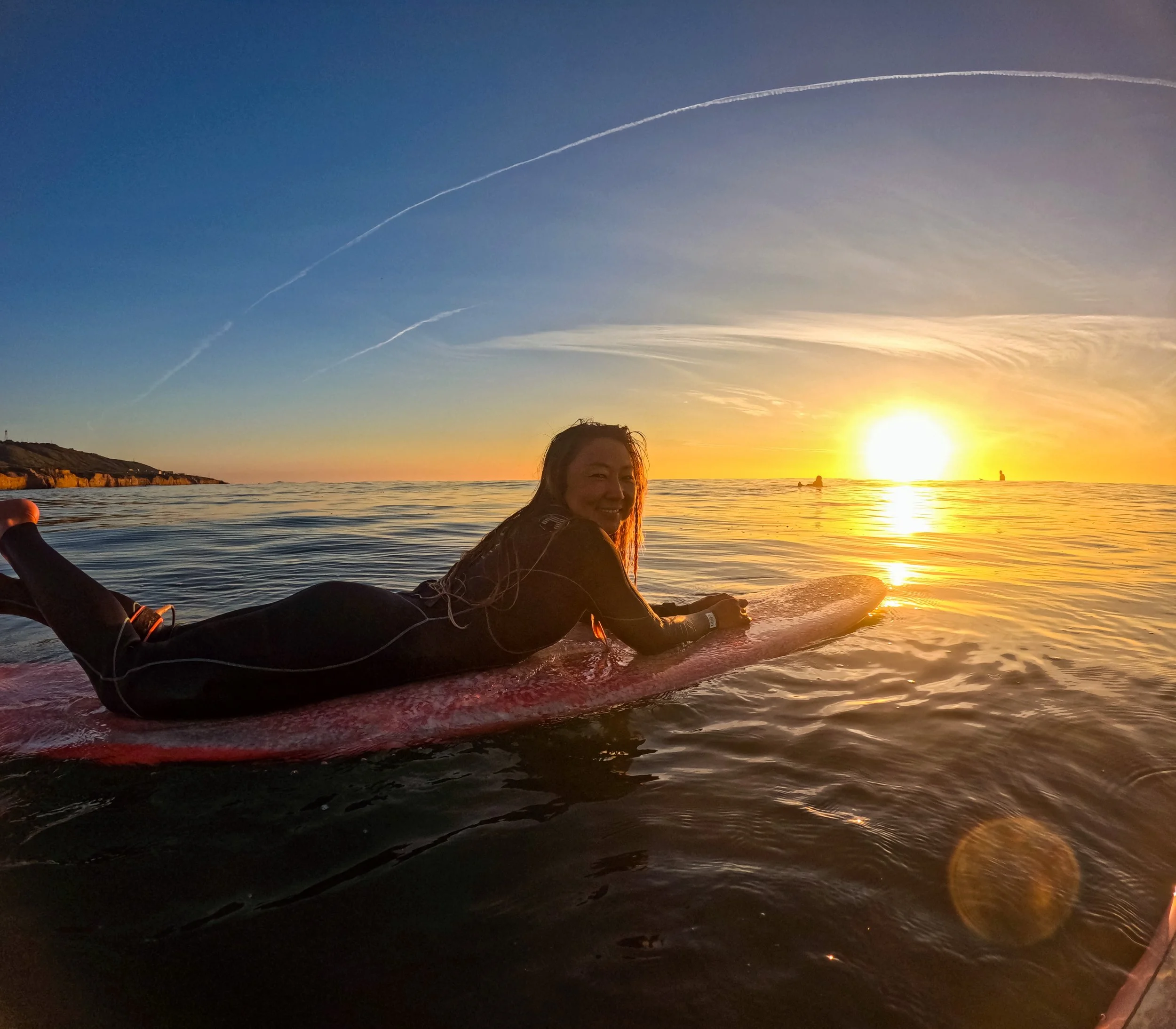 Woman lying on a surfboard in the ocean at sunset, smiling with the sun setting on the horizon and boats in the distance.