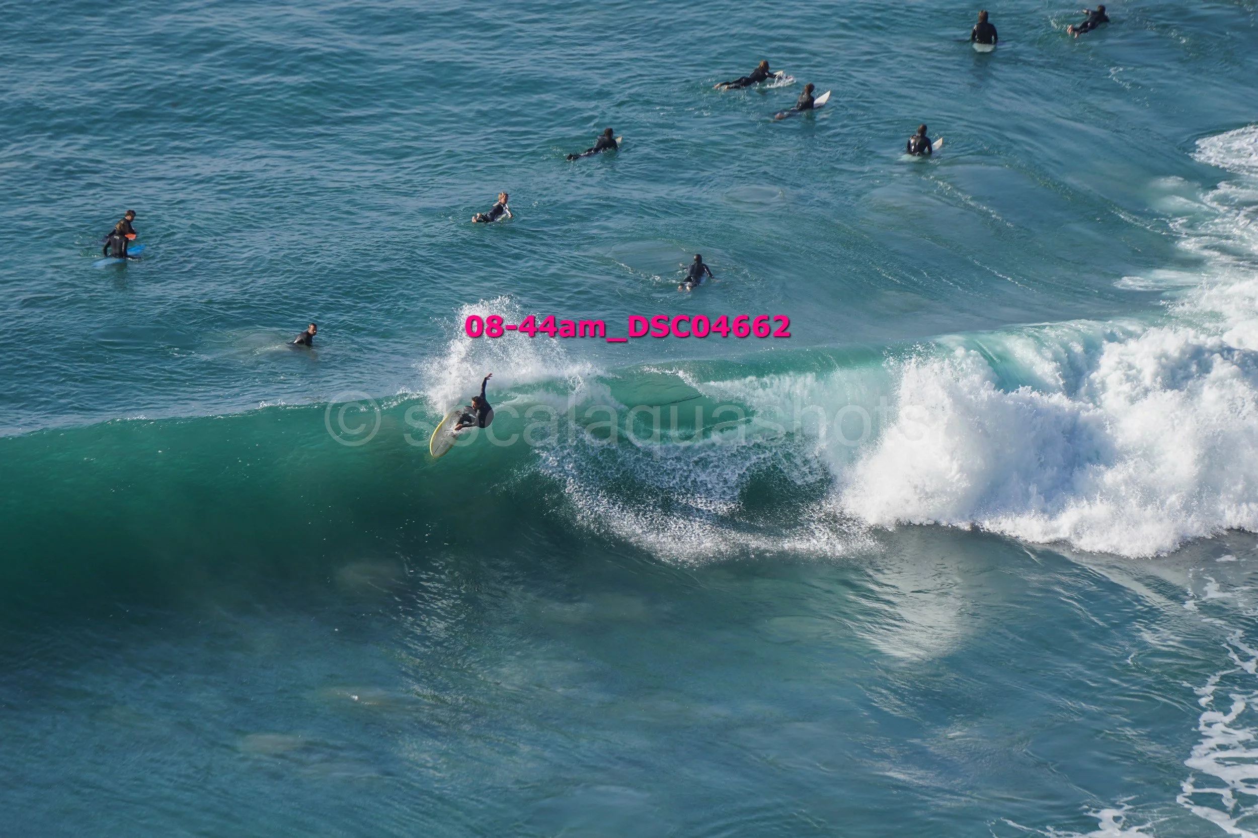 Surfer riding a large wave with many people in wetsuits swimming or floating in the water nearby.