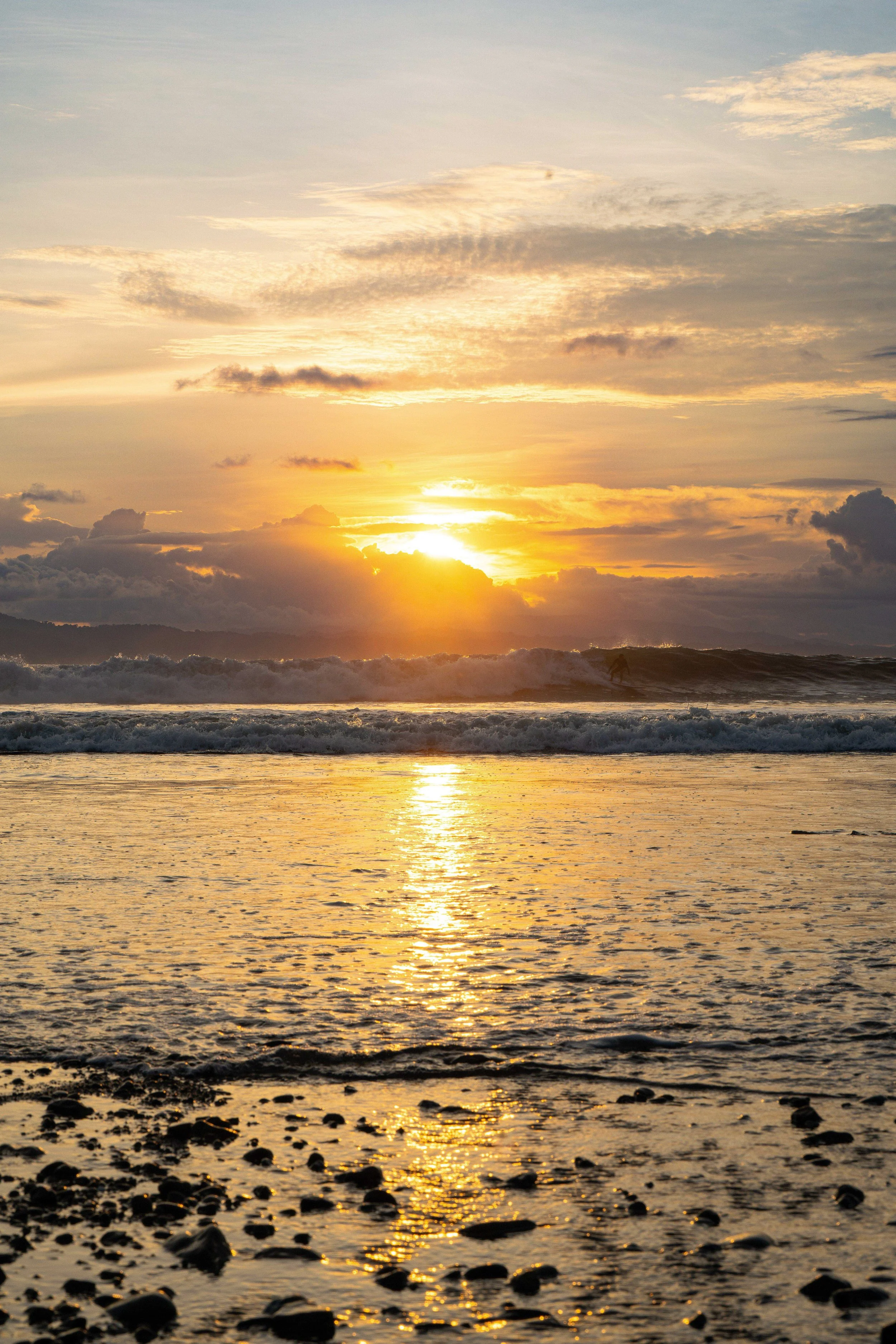 Sunset over the ocean with clouds and waves crashing on the shore.