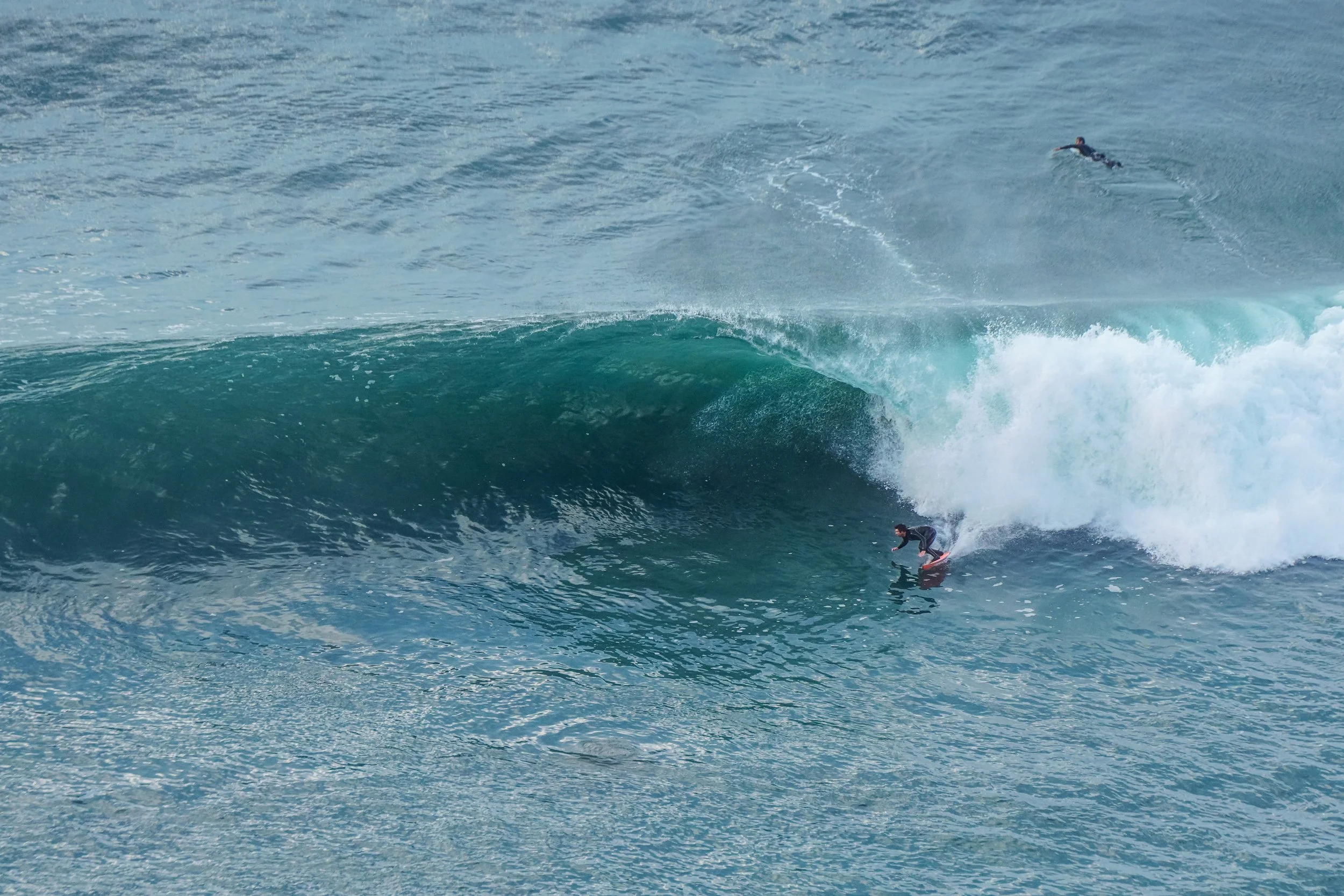 A surfer riding a large wave in the ocean, with another surfer swimming in the background.