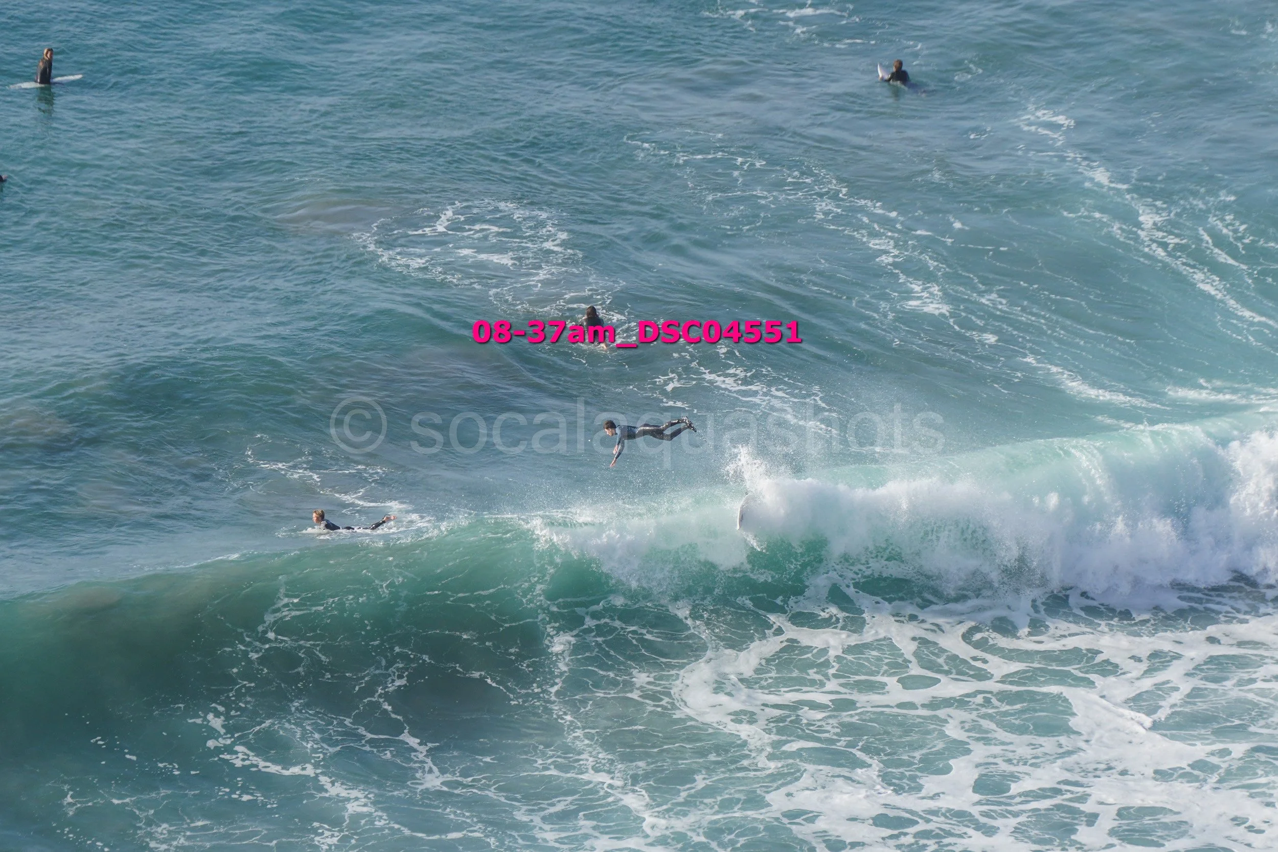 Multiple surfers on the ocean, with some falling off their surfboards and others paddling on their boards amidst large crashing waves