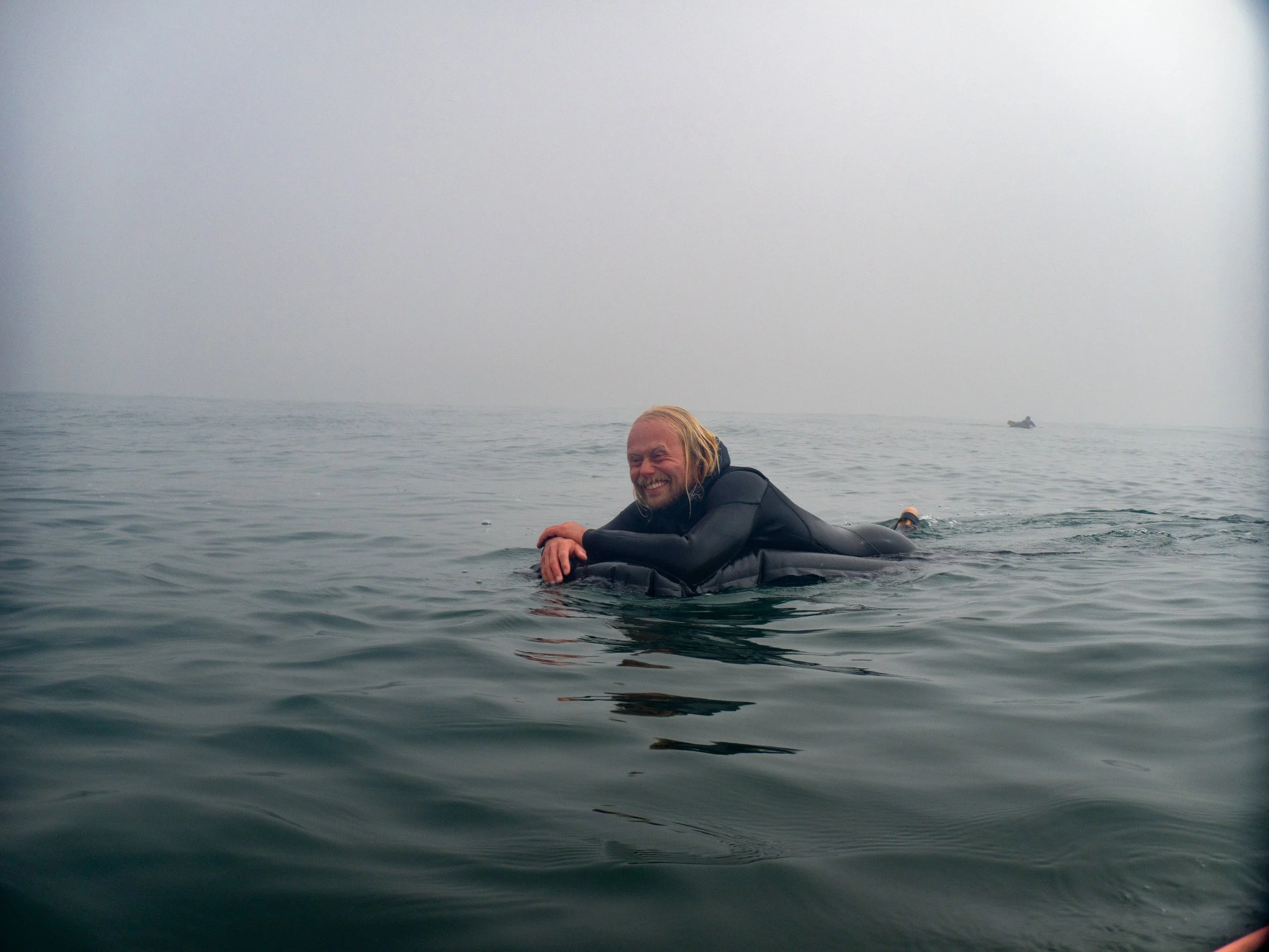 A person is lying on a surfboard in the ocean, smiling, with foggy weather and another small boat in the distance.