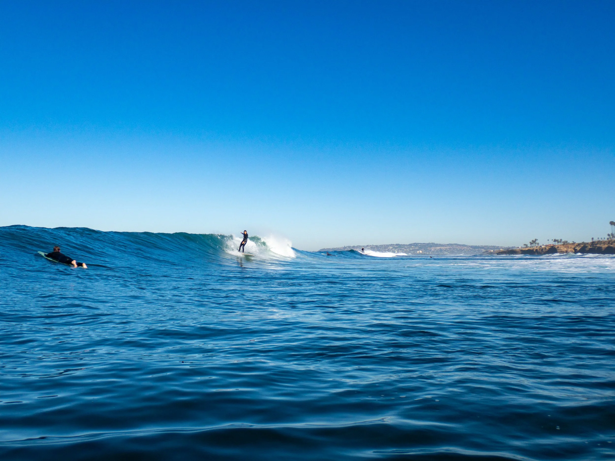 A person surfing a wave while another person paddles in the water, with a coastline and palm trees in the background under a clear blue sky.