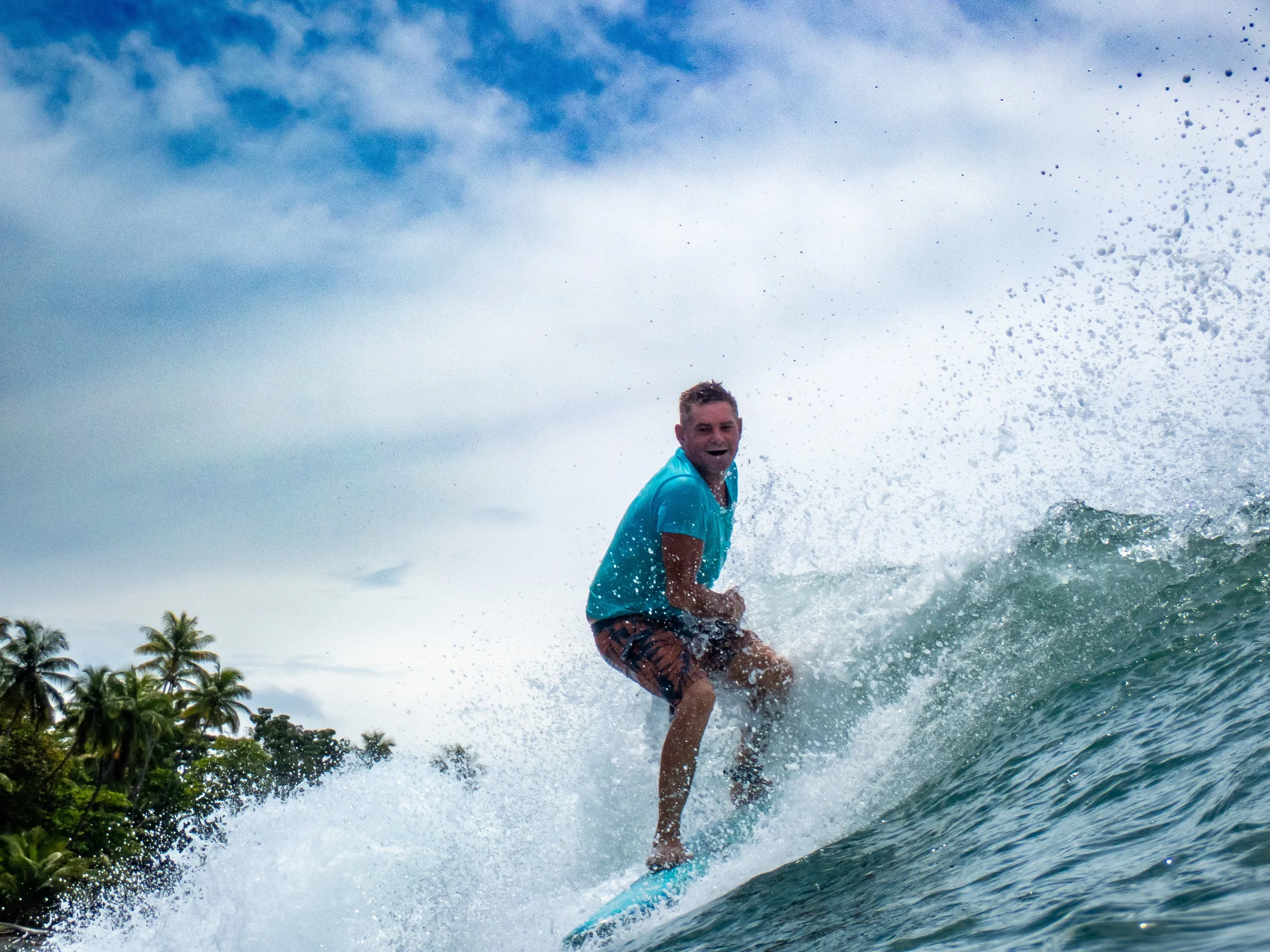 A man surfing on a wave in the ocean, wearing a blue shirt and shorts, smiling, with palm trees and a cloudy sky in the background.
