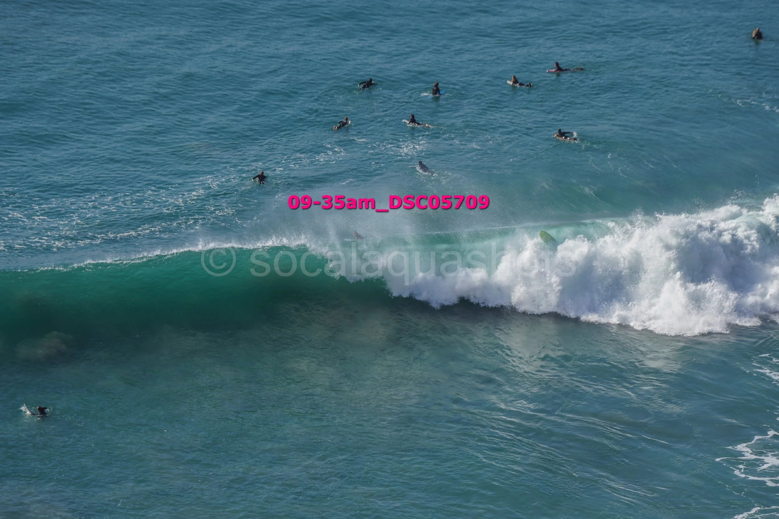 Multiple surfers in the water, some on surfboards riding a wave, others waiting. The ocean is a deep blue-green with white foamy waves.