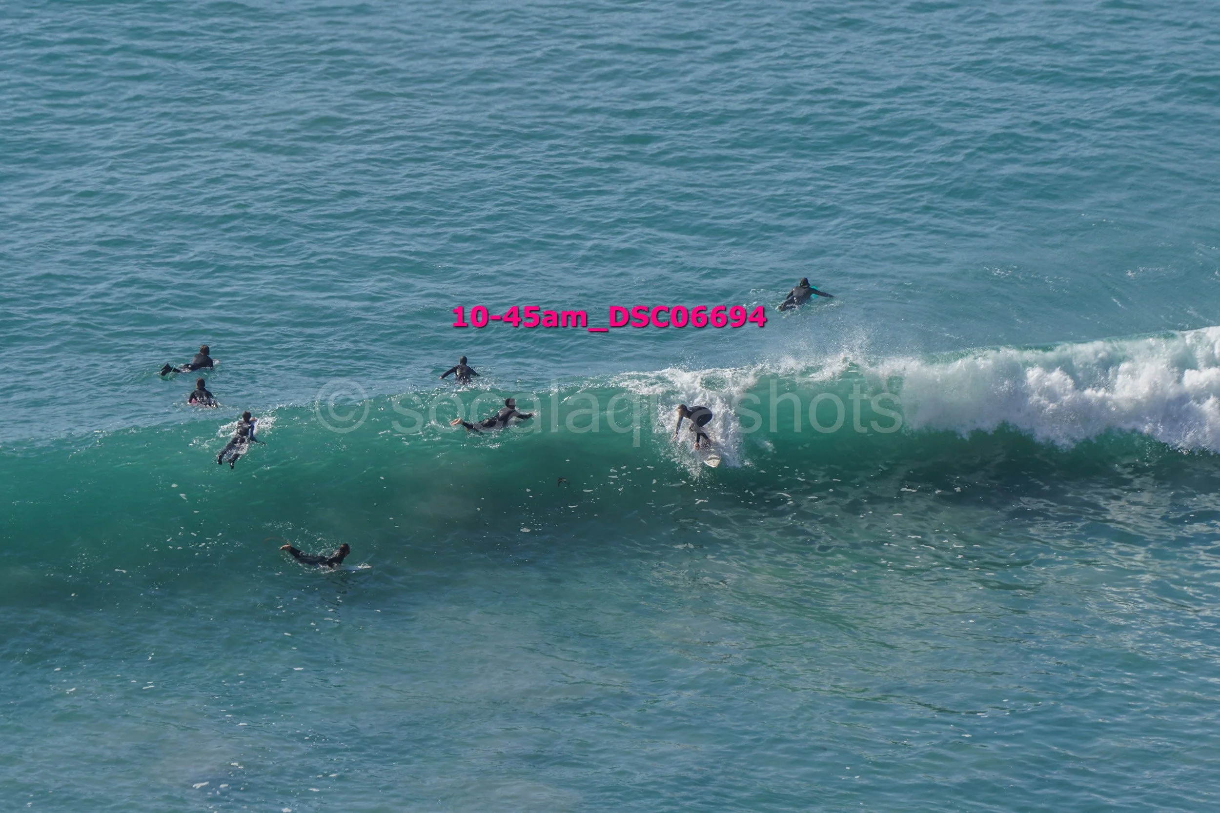 Group of surfers riding a wave in the ocean with clear blue water, some paddling and some riding the wave.