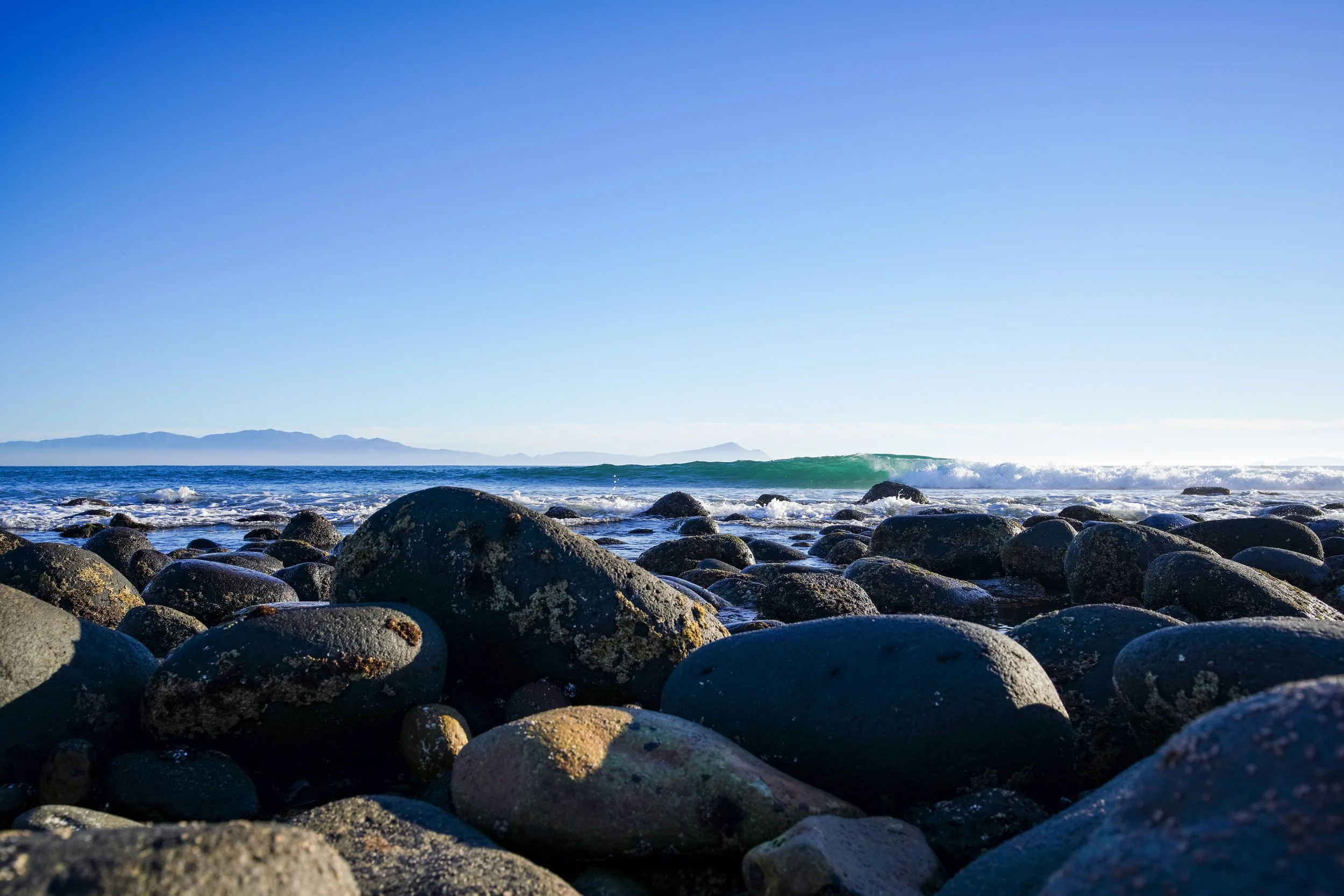 A rocky beach scene with smooth stones in the foreground and ocean waves crashing against rocks, under a clear blue sky with distant mountains in the background.
