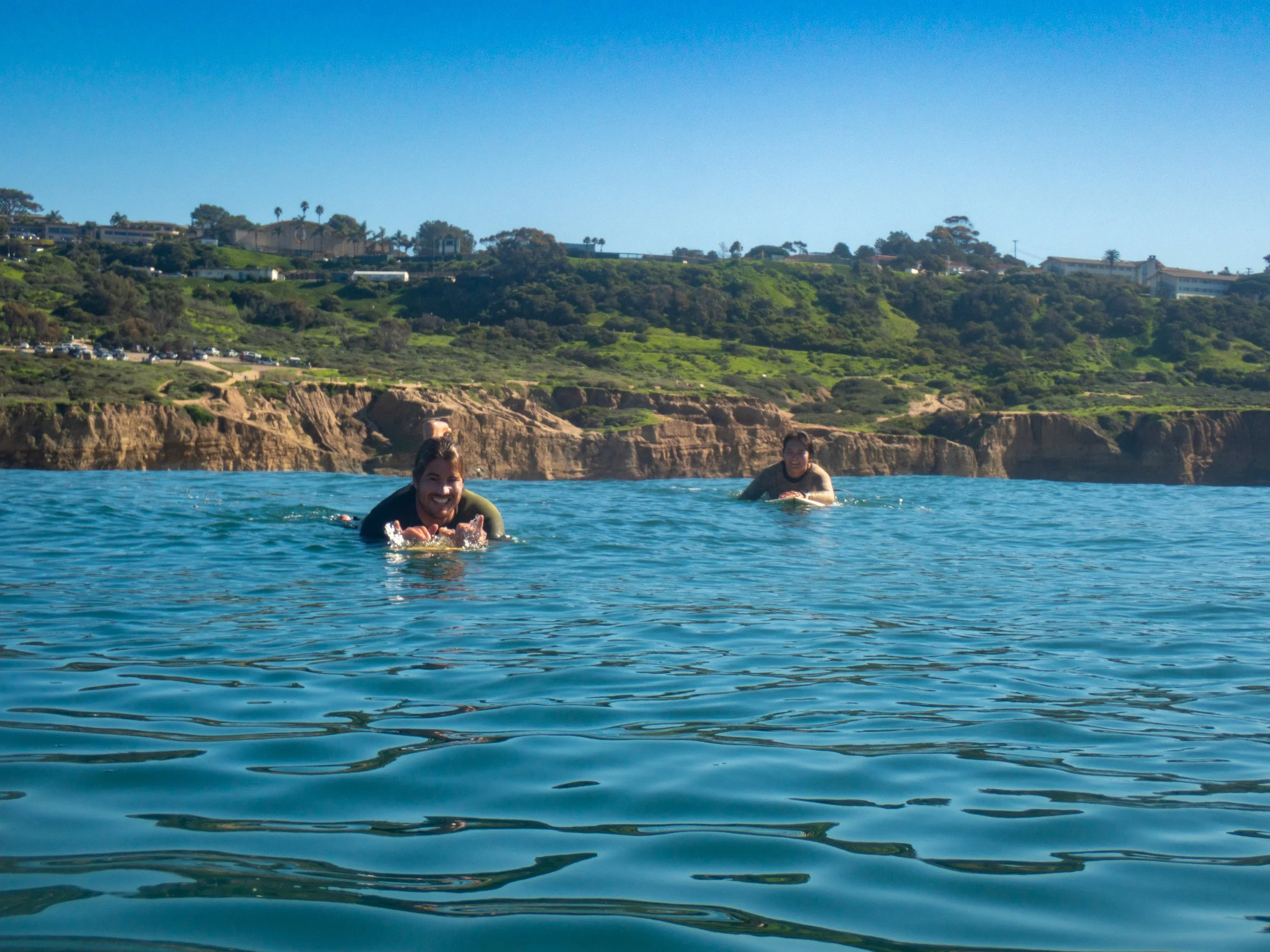 Two women swimming in the ocean near the coast, with green hills, cliffs, and houses in the background under a clear blue sky.