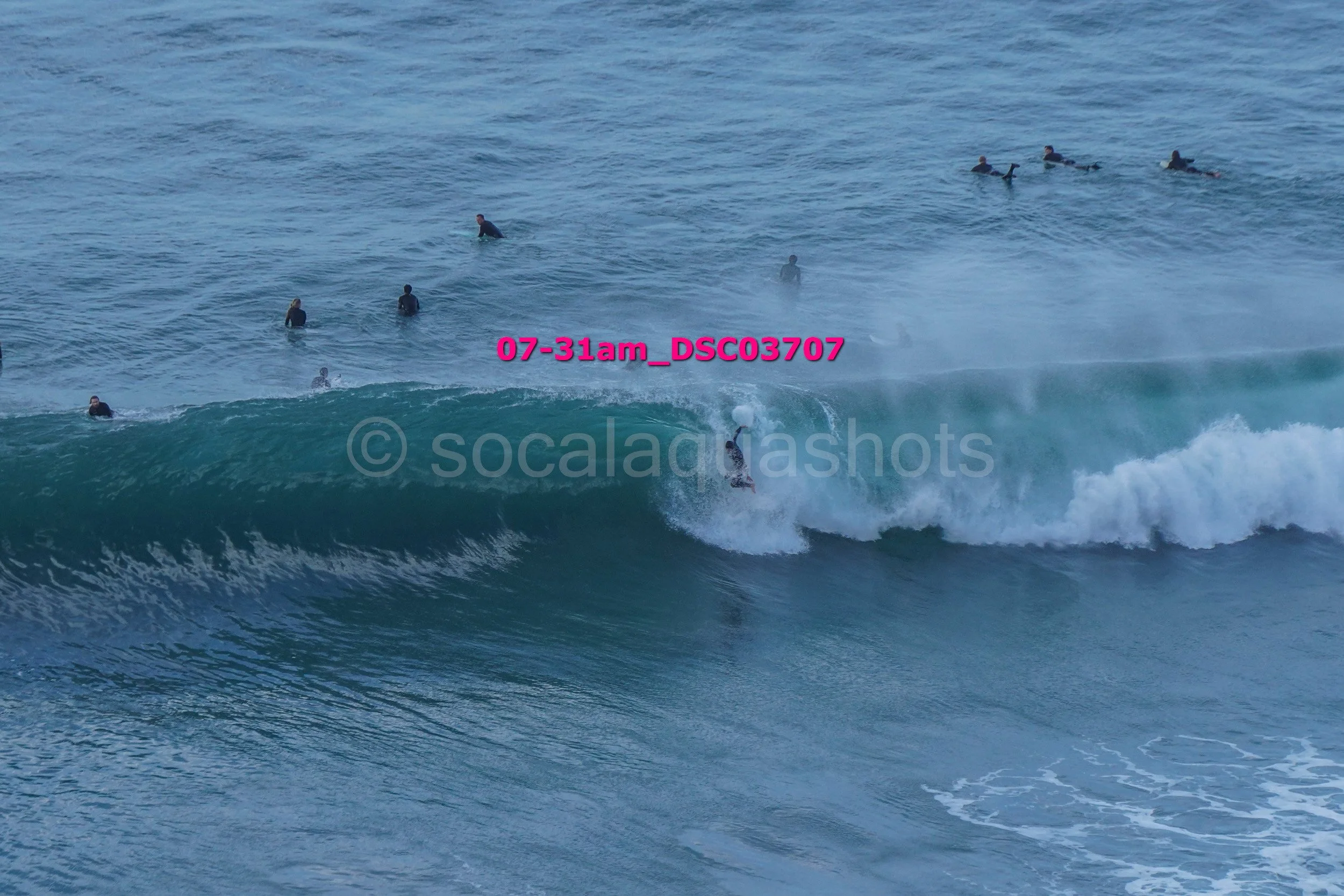 A group of surfers in the ocean, with one surfer riding a wave and others waiting or swimming in the water.