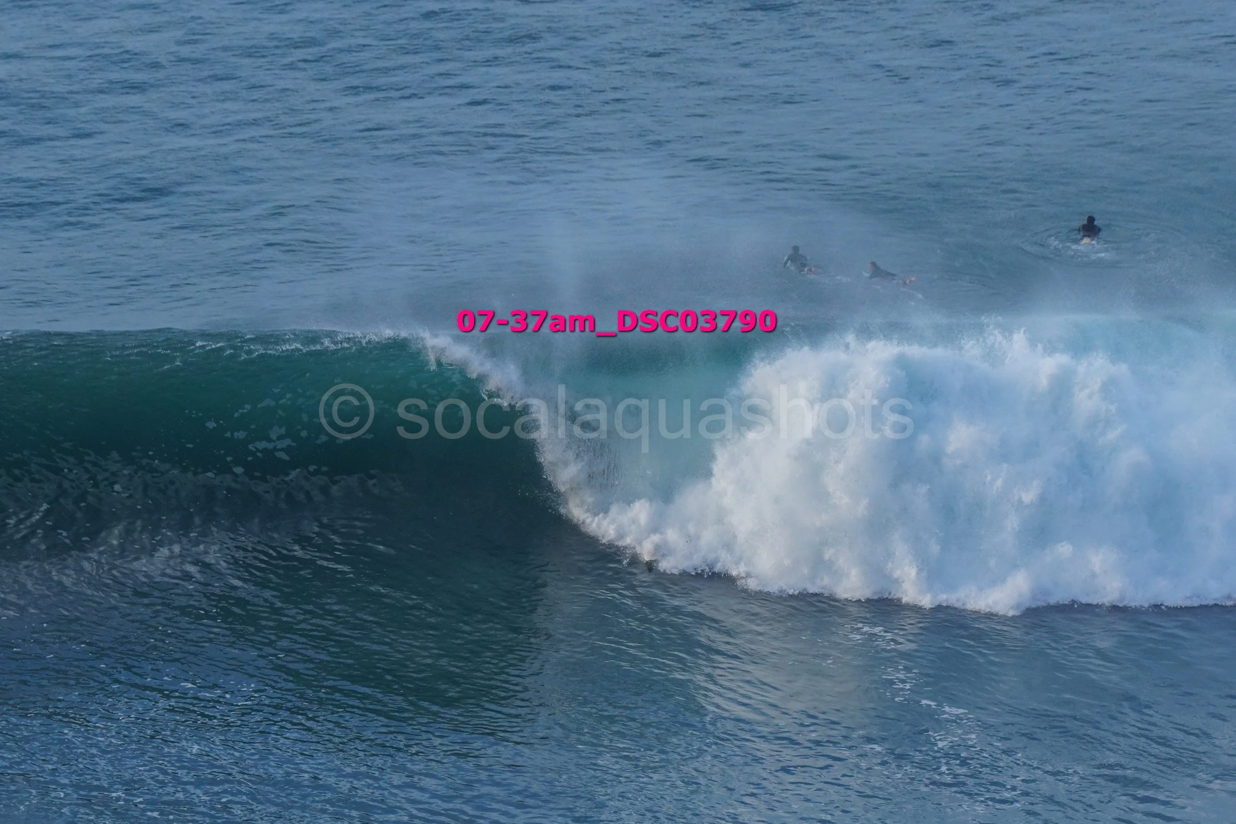 A wave breaking in the ocean with three surfers in the background.