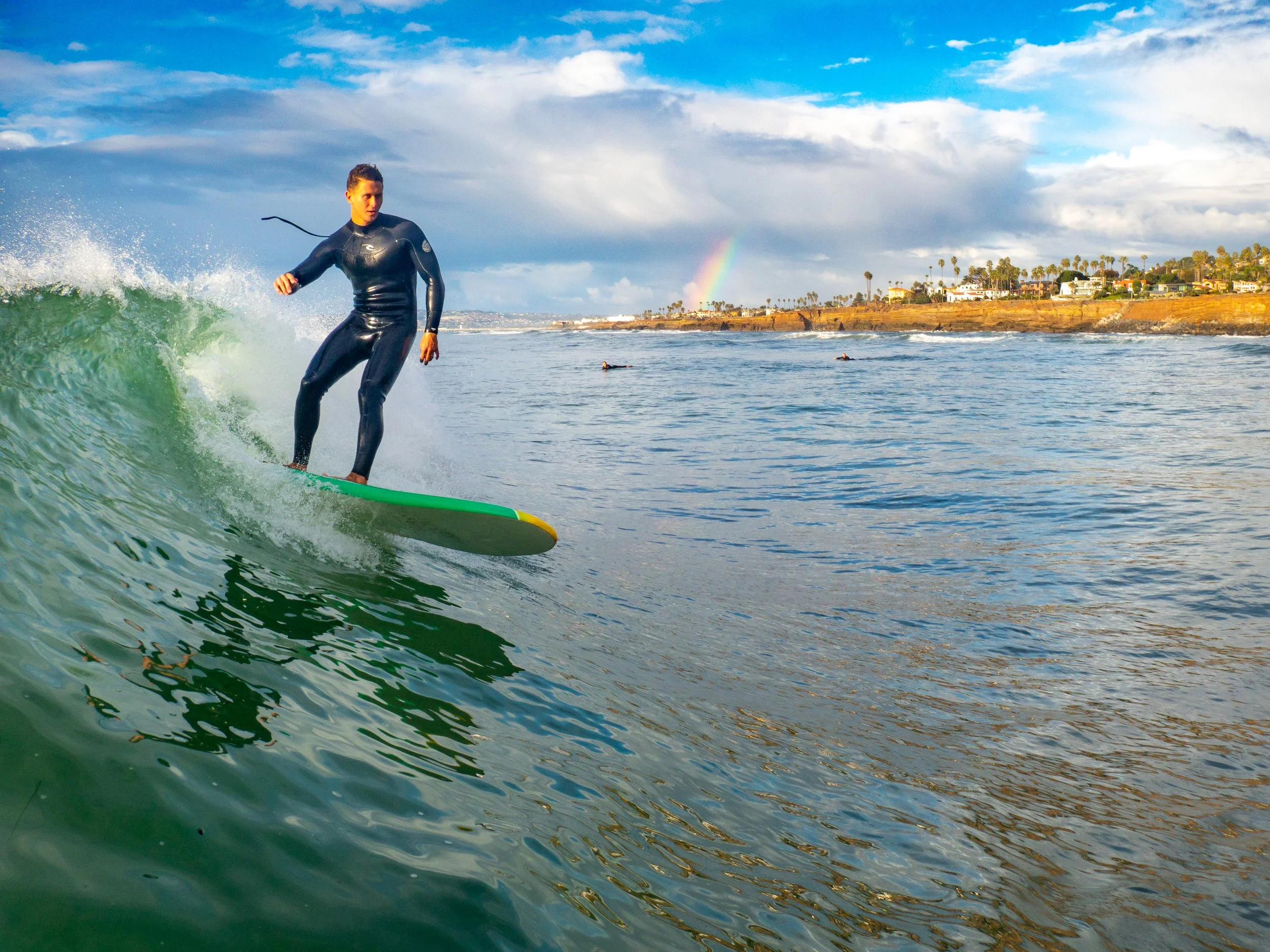 Woman in black wetsuit surfing on a green surfboard near the shoreline with a rainbow and houses in the background