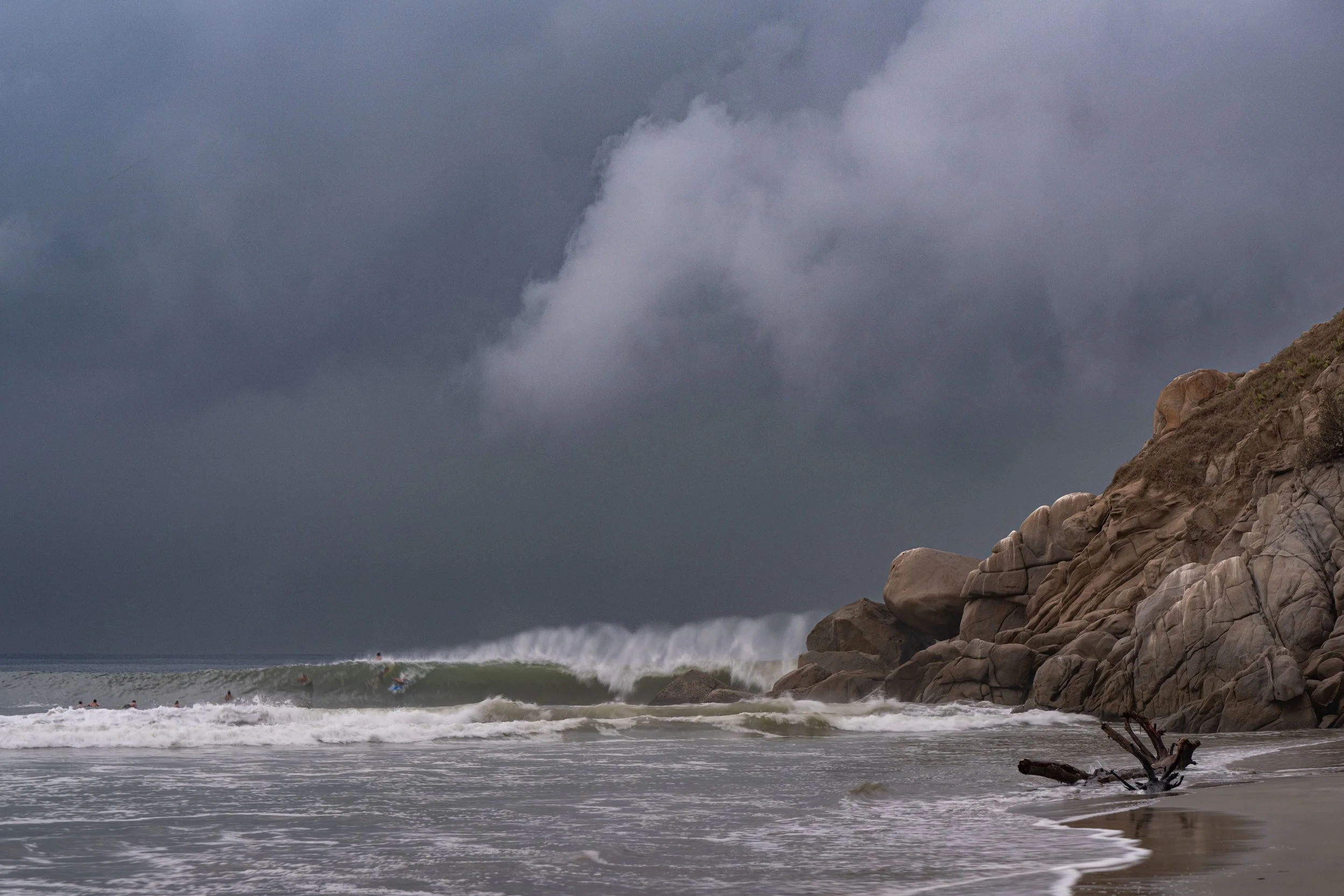 A cloudy sky over the ocean with waves breaking near a rocky shoreline and surfers in the water.