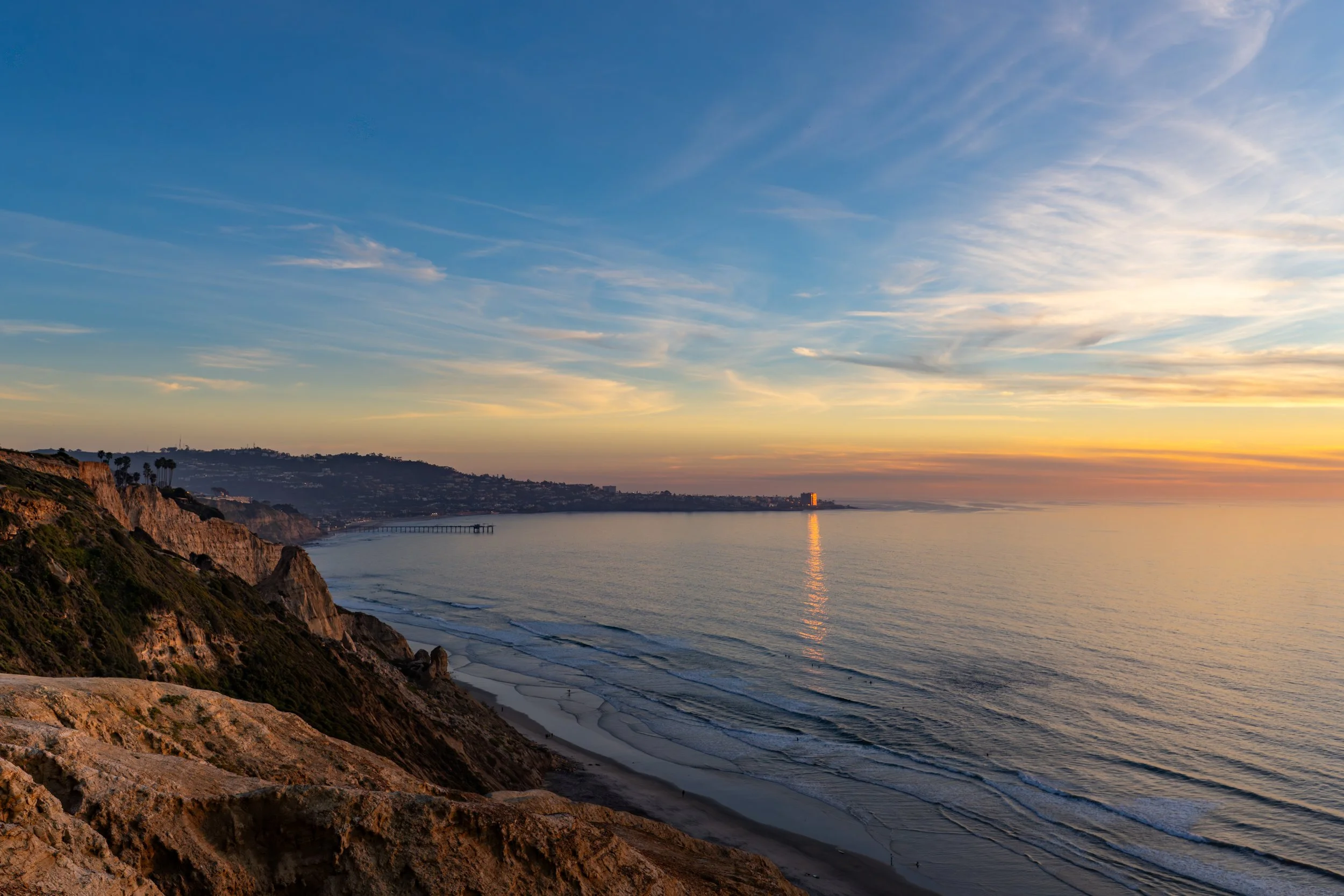 Coastal cliffs overlooking an ocean at sunset with a pier extending into the water and a distant city skyline.