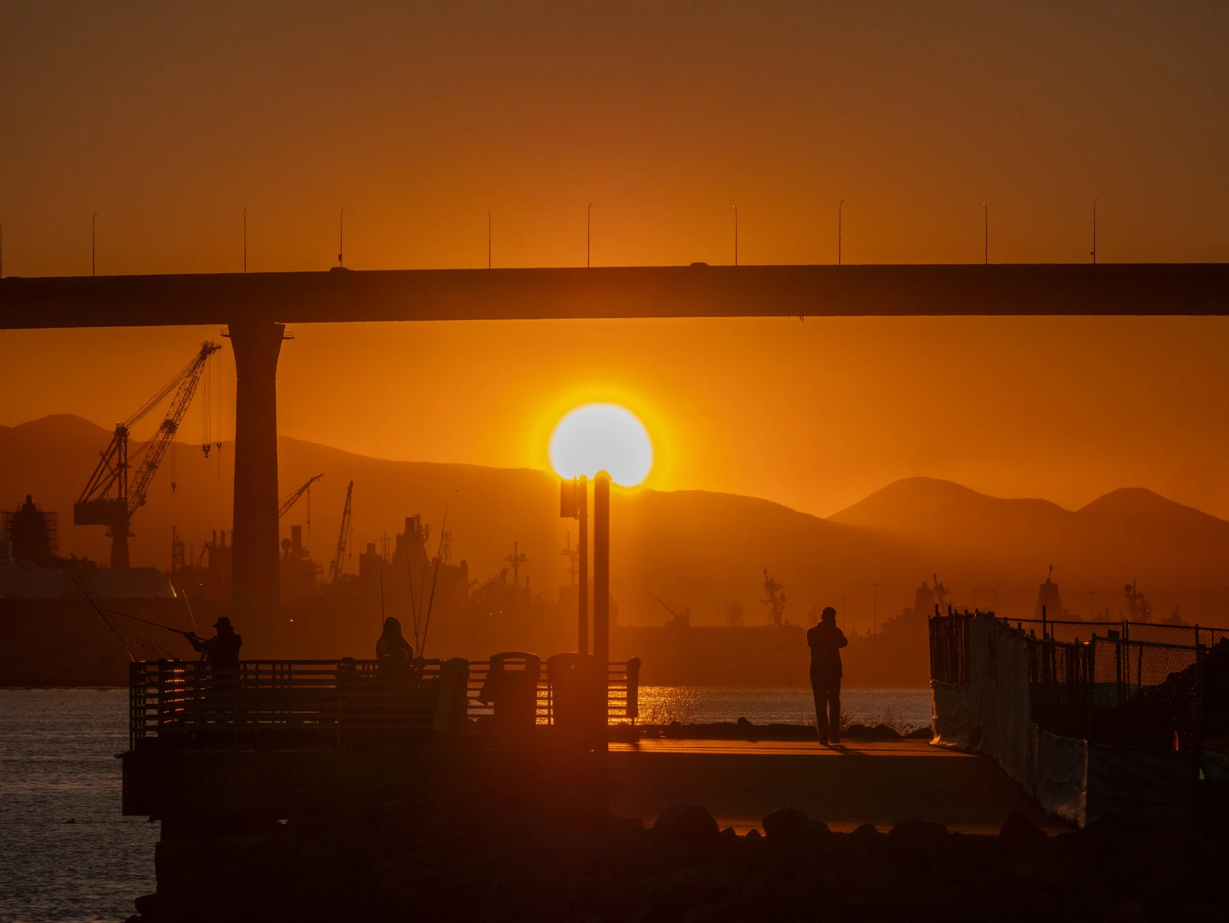 Silhouettes of fishermen on a dock at sunset, with a bridge and mountains in the background, and construction cranes in a harbor.