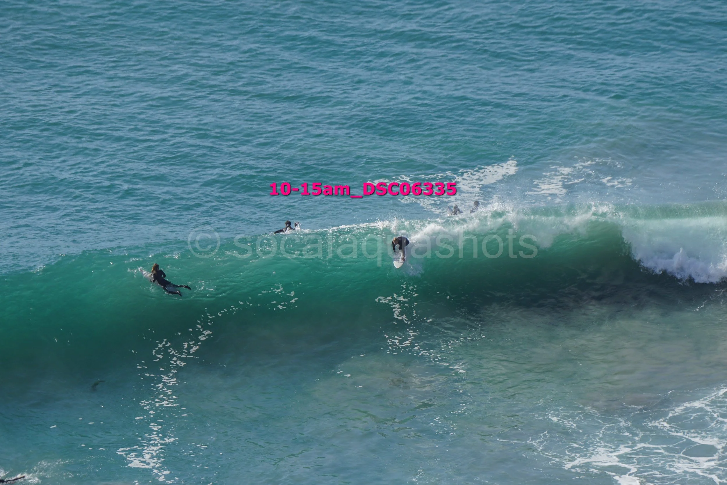 Multiple surfers riding and paddling on a large ocean wave with a group of surfers in the water nearby.