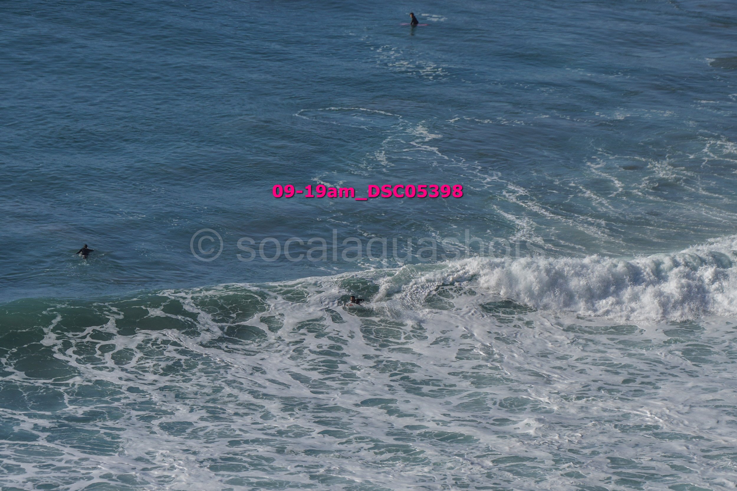 Two surfers in the ocean, one paddling on the left and another partly submerged near white waves, with a person surfing in the distance.