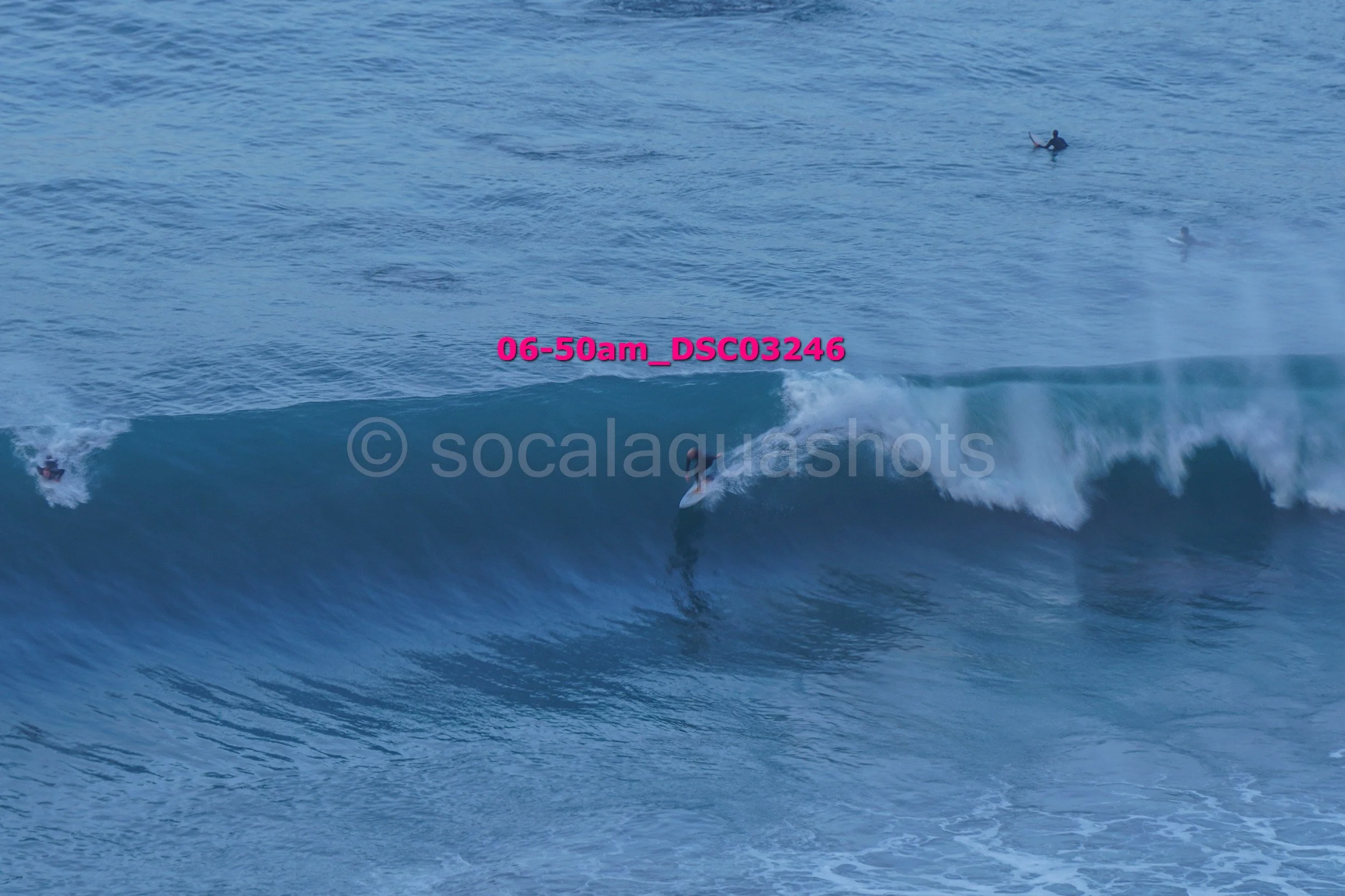 A person surfing on a wave in the ocean with two other surfers in the background and the timestamp '06-50am DSC03246' in pink text across the image.