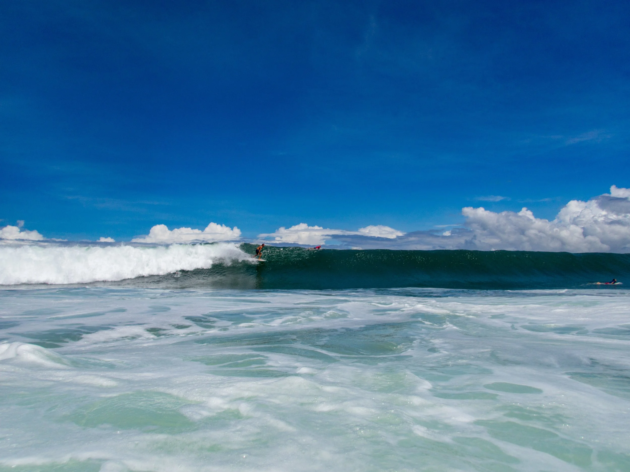 Surfer riding a large ocean wave under a blue sky with clouds.