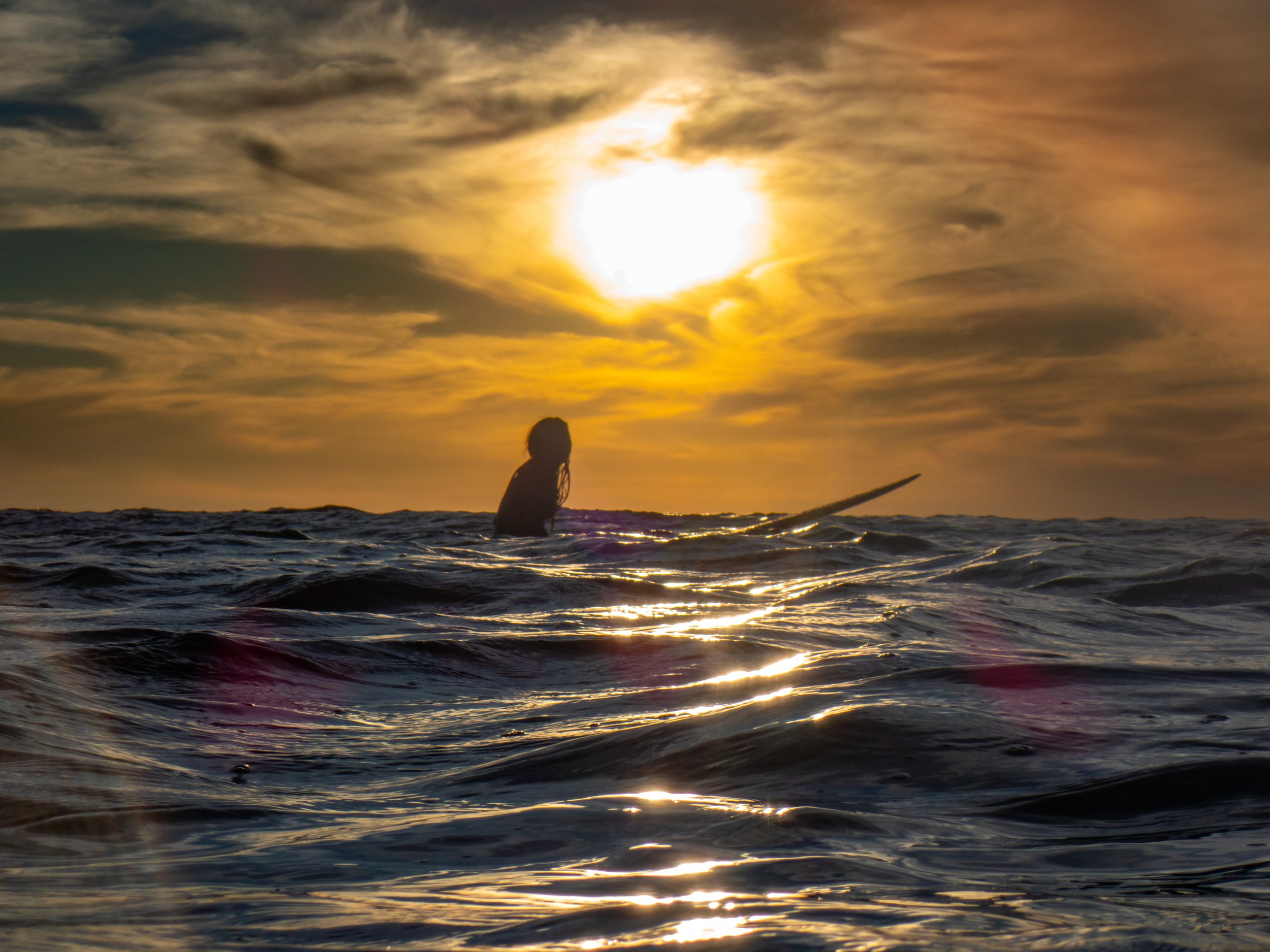 Silhouette of a person holding a surfboard in the ocean during sunset with a partly cloudy sky.