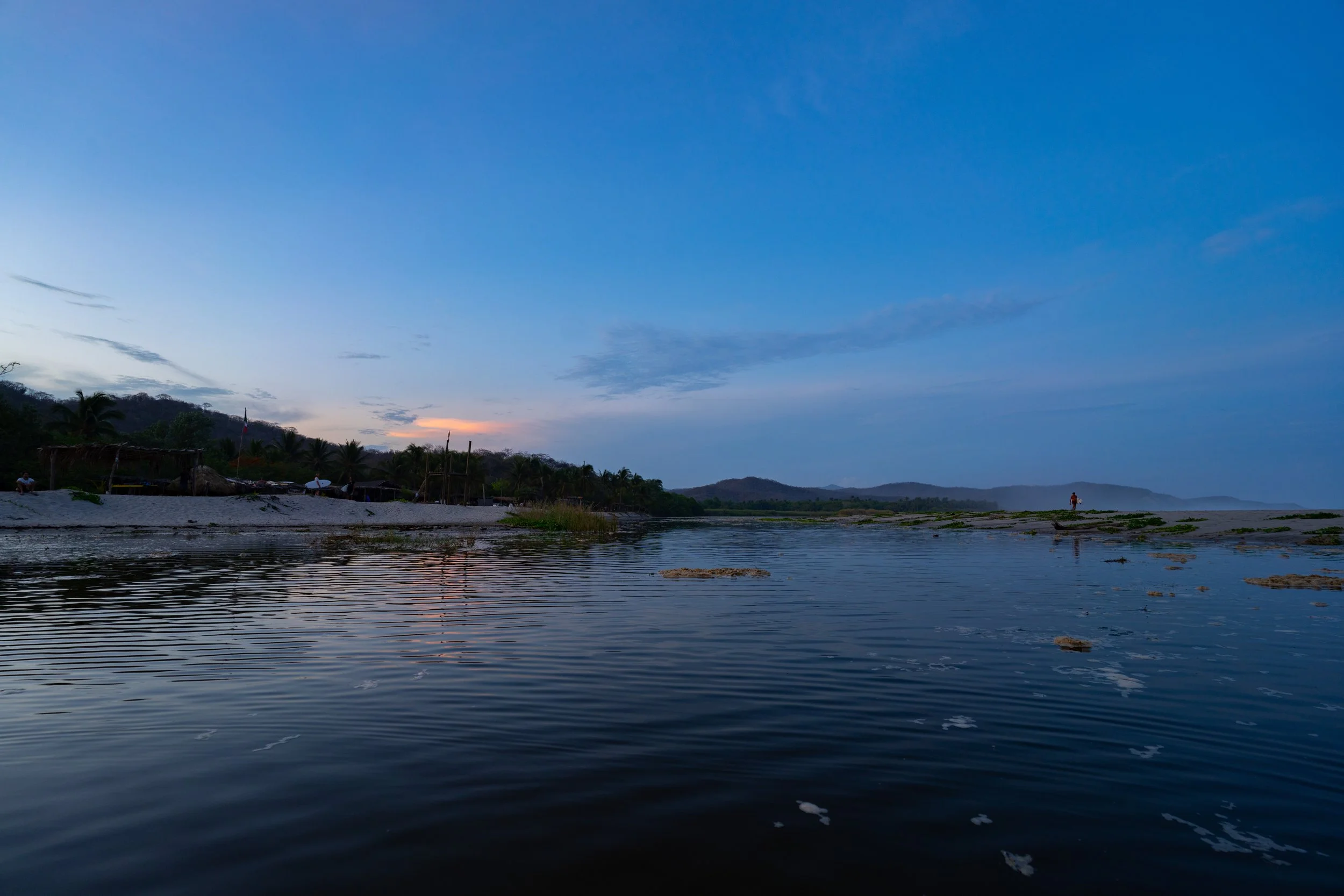 Sunset over a calm river with a distant shoreline, small huts, and trees, under a partly cloudy sky.