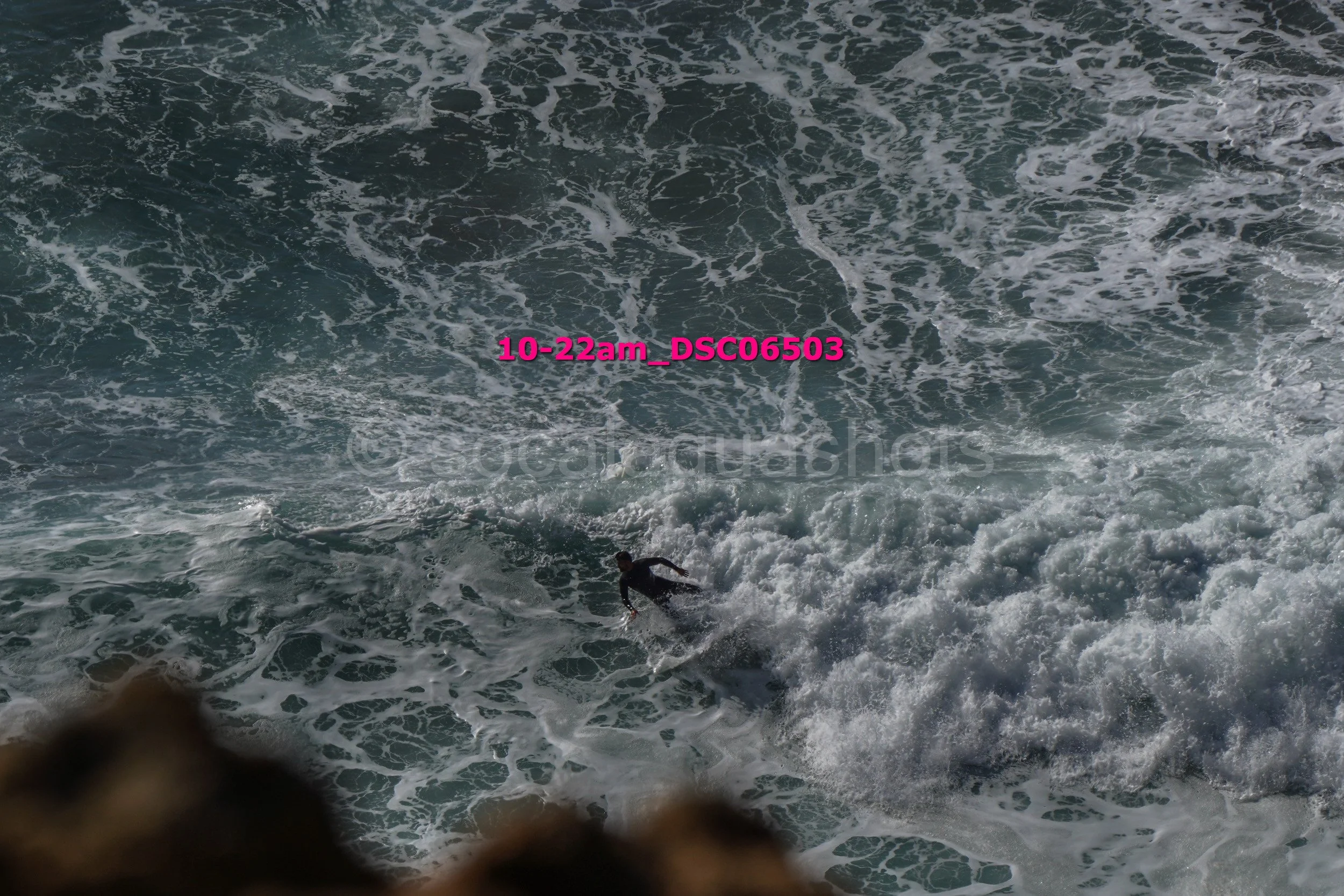 A person surfing in rough ocean waves, viewed from above with rocks at the bottom of the image.