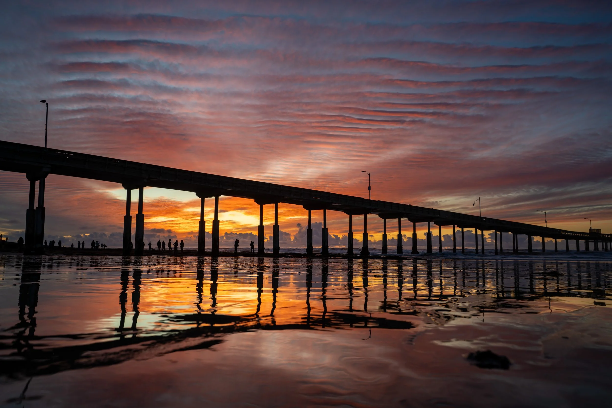 Sunset over a pier with silhouetted people walking along the beach, with vibrant pink, orange, and purple clouds reflected on the wet sand