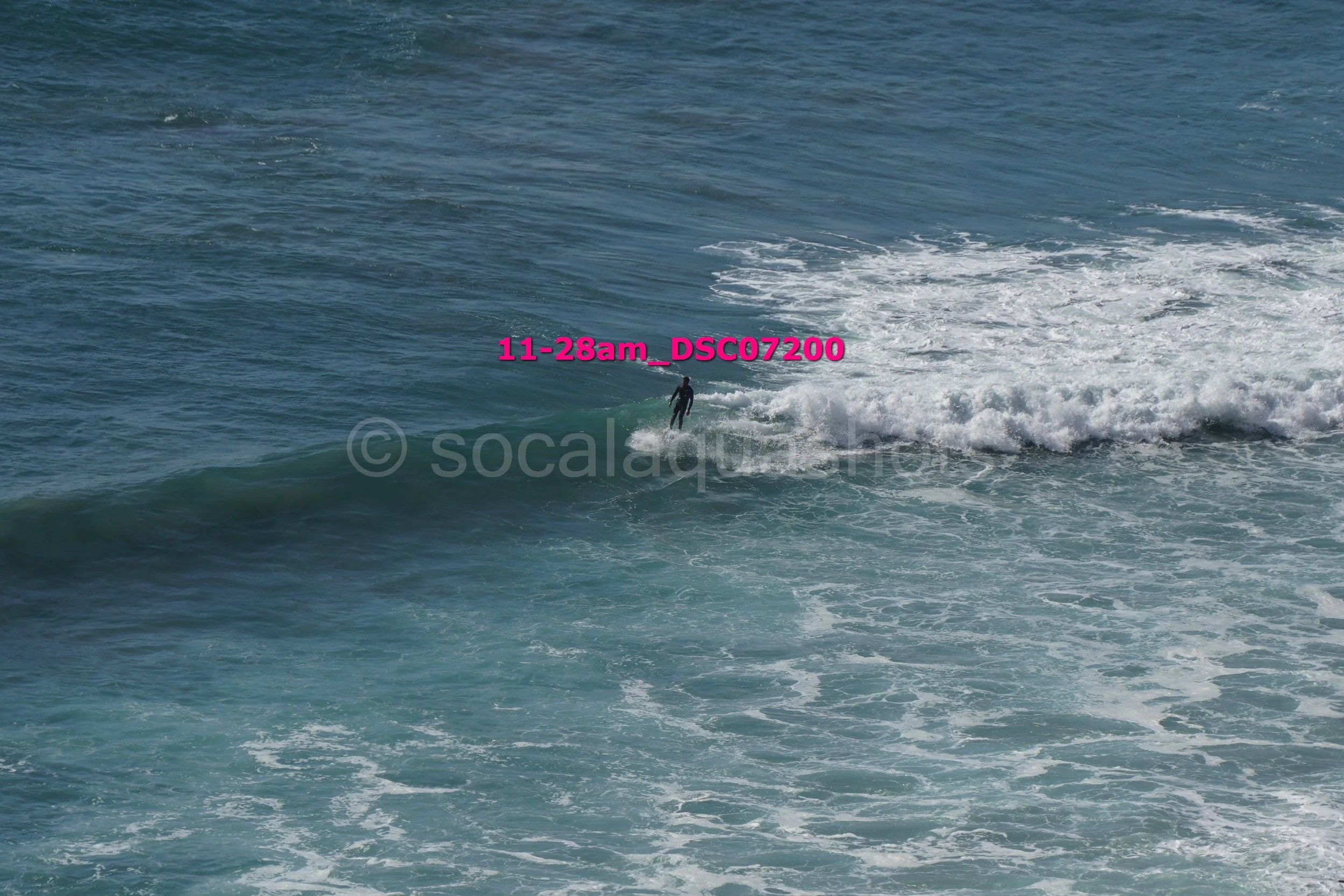 A surfer riding a wave in the ocean visible from afar with a pink timestamp overlay reading '11-28am_DSC07200'.