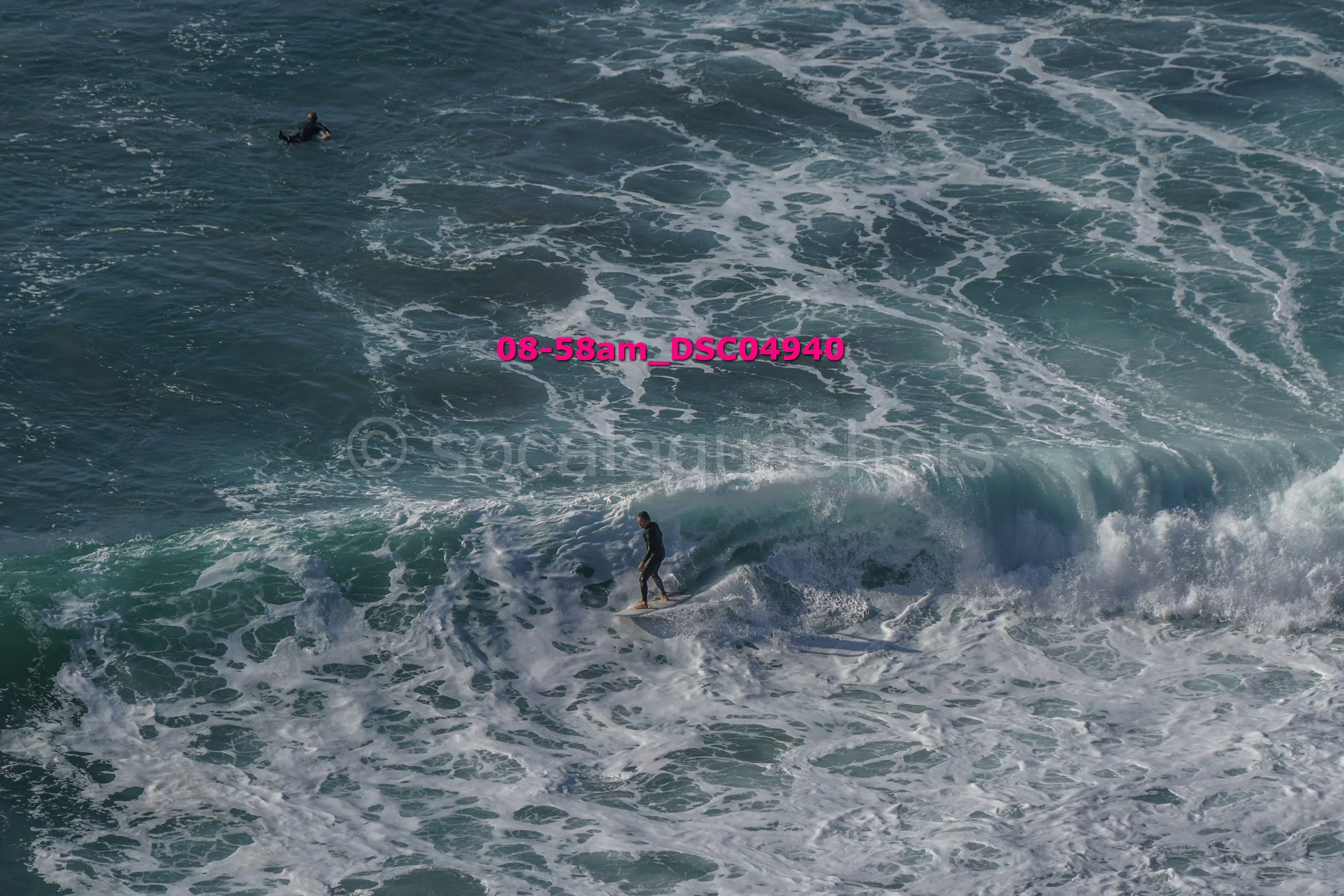 A person surfing on a wave in the ocean, with another surfer visible in the distance.
