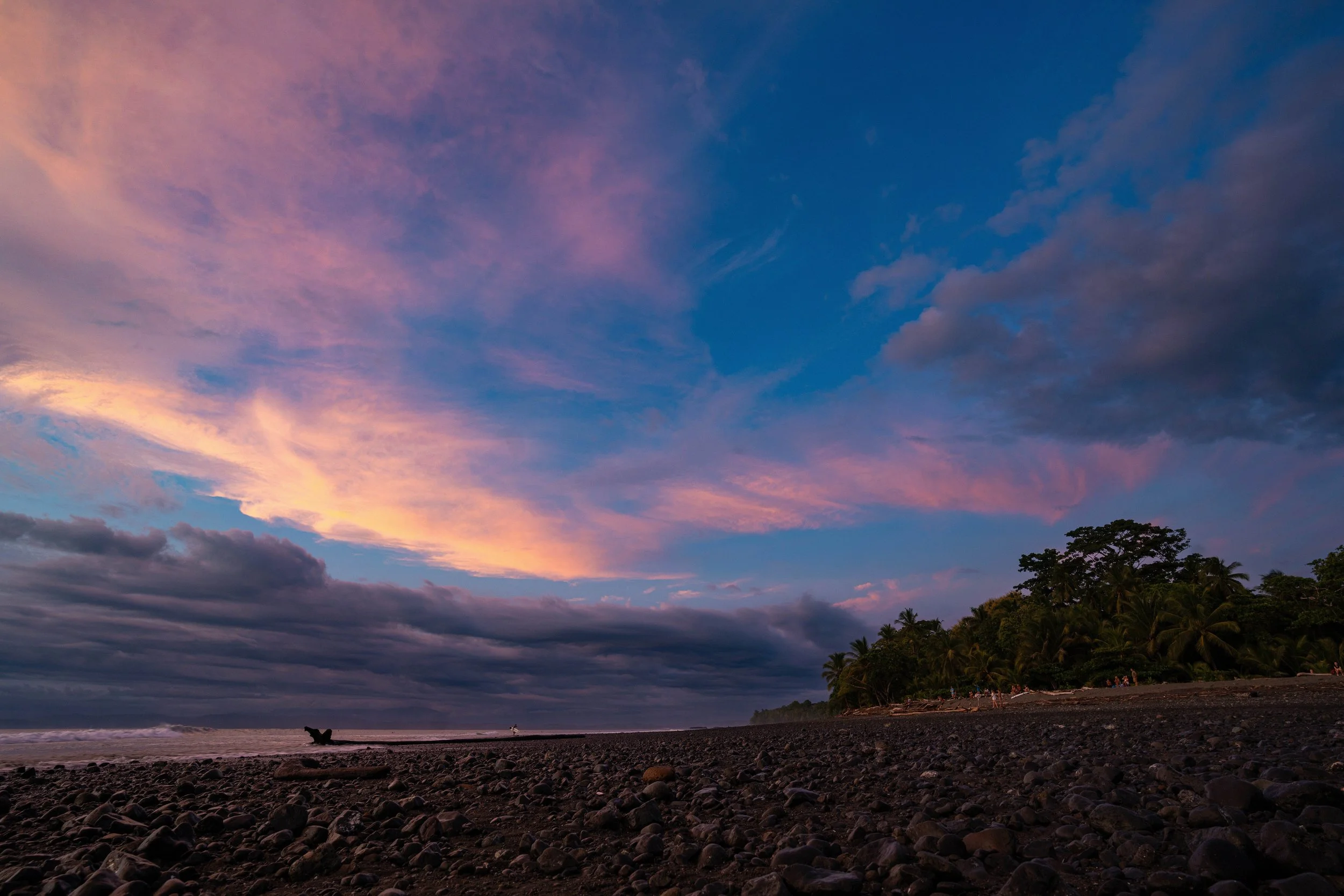 A colorful sky during sunset over a rocky beach with trees in the distance.