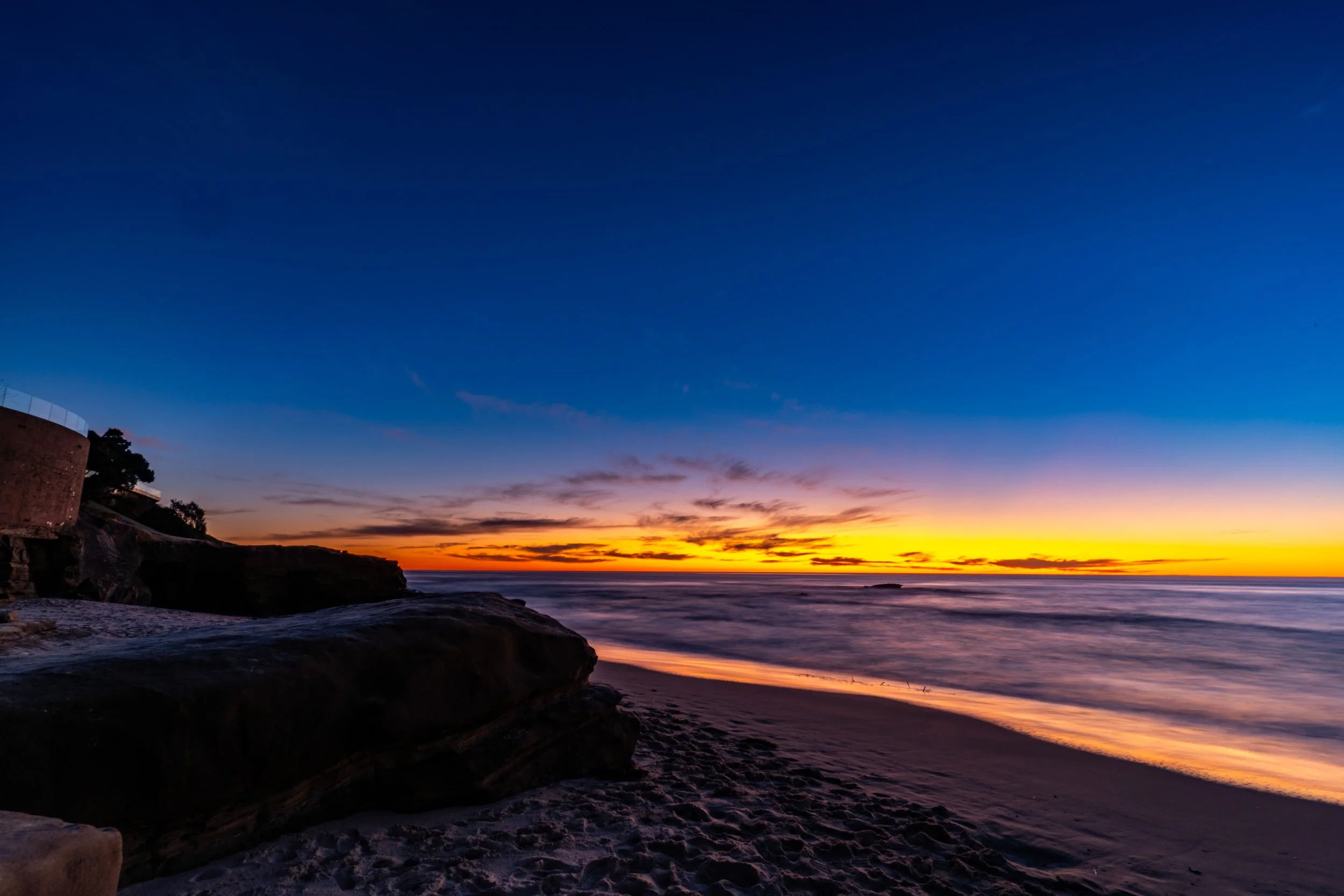 Sunset over the ocean with orange and yellow colors near the horizon, dark rocks and sandy beach in the foreground, and a clear evening sky changing from orange to deep blue.