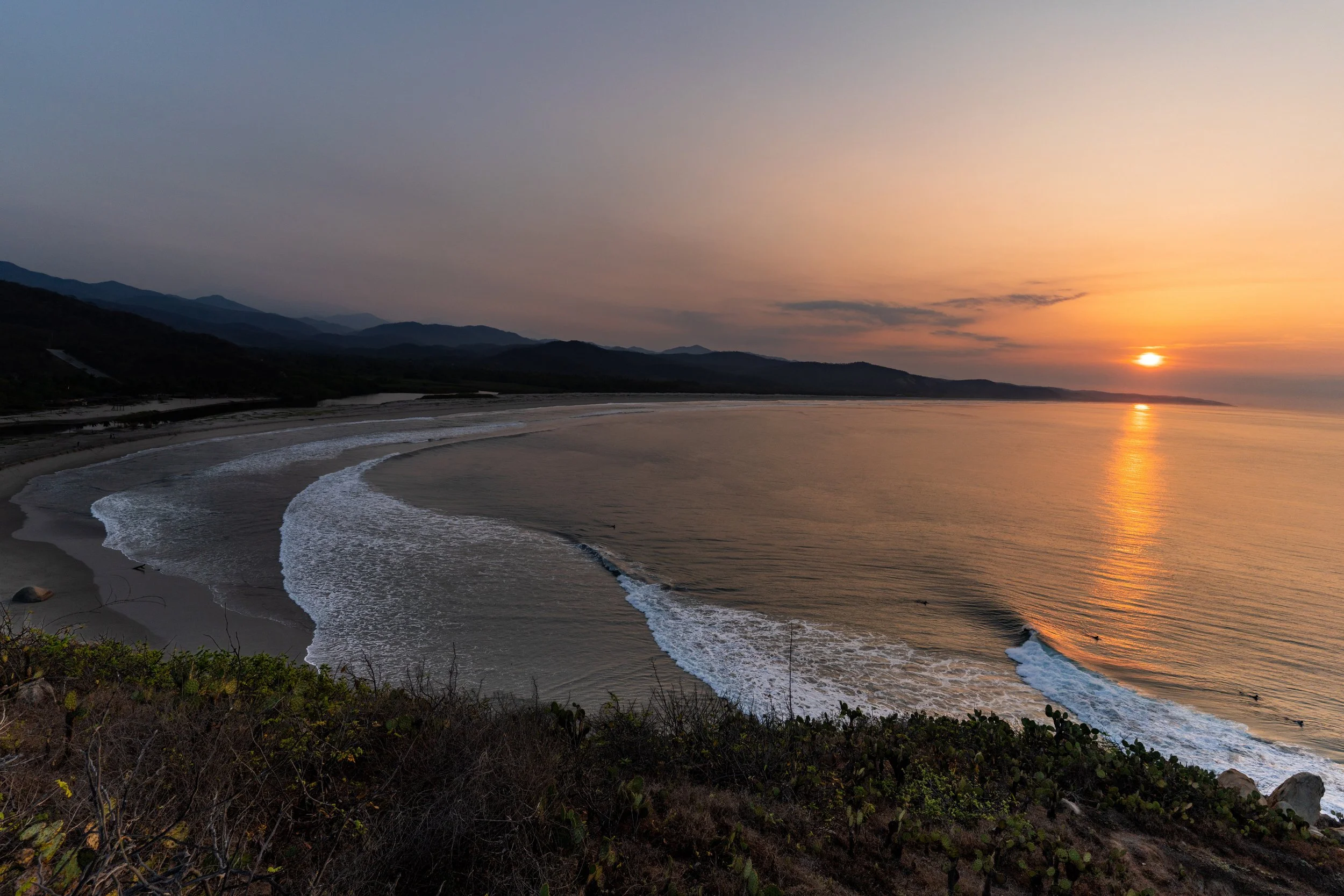Beach at sunset with waves crashing on shore, mountains in background, and the sun near the horizon.