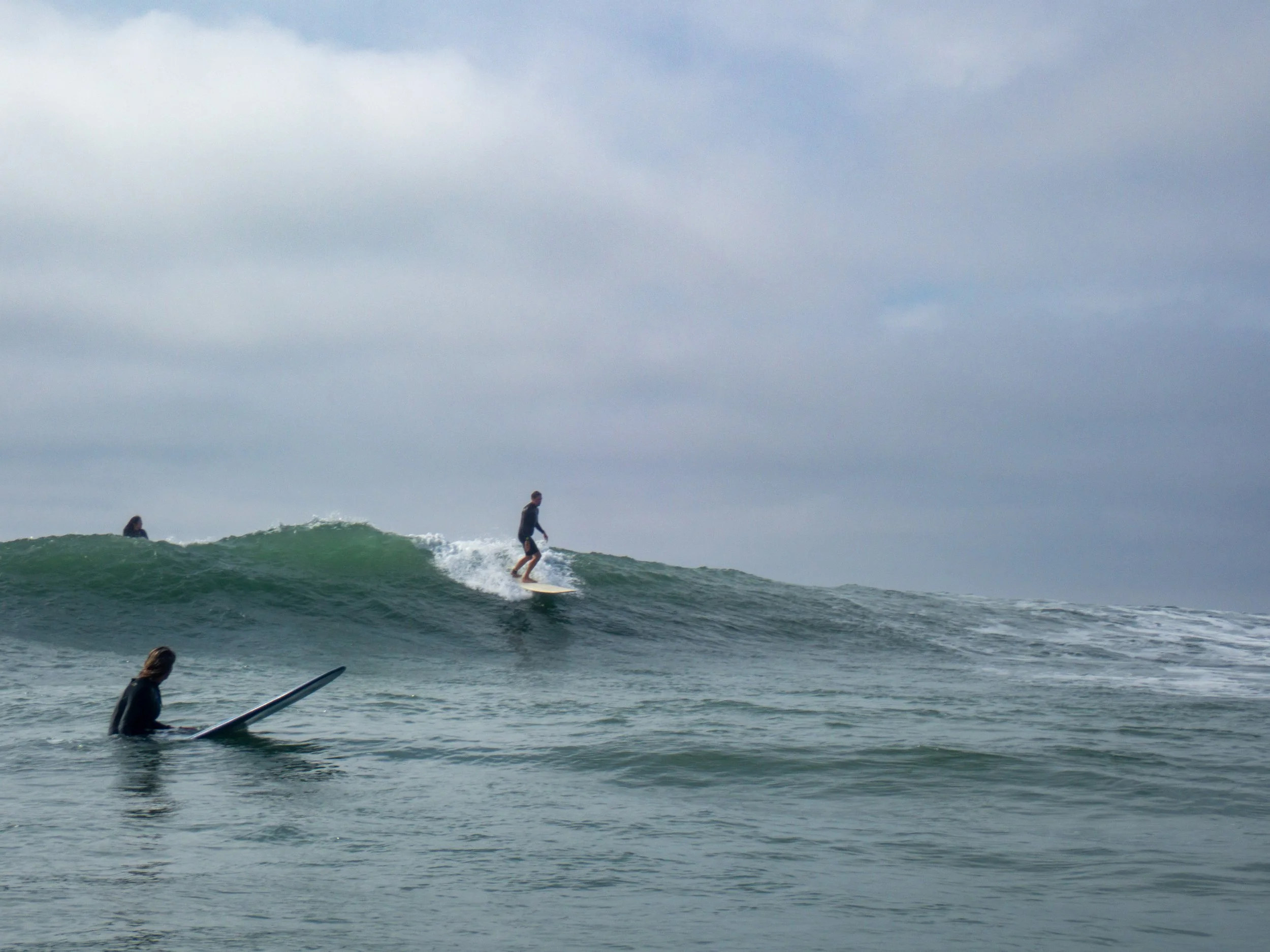 A surfer riding a wave on a surfboard in the ocean, with other surfers in the distance and overcast sky.