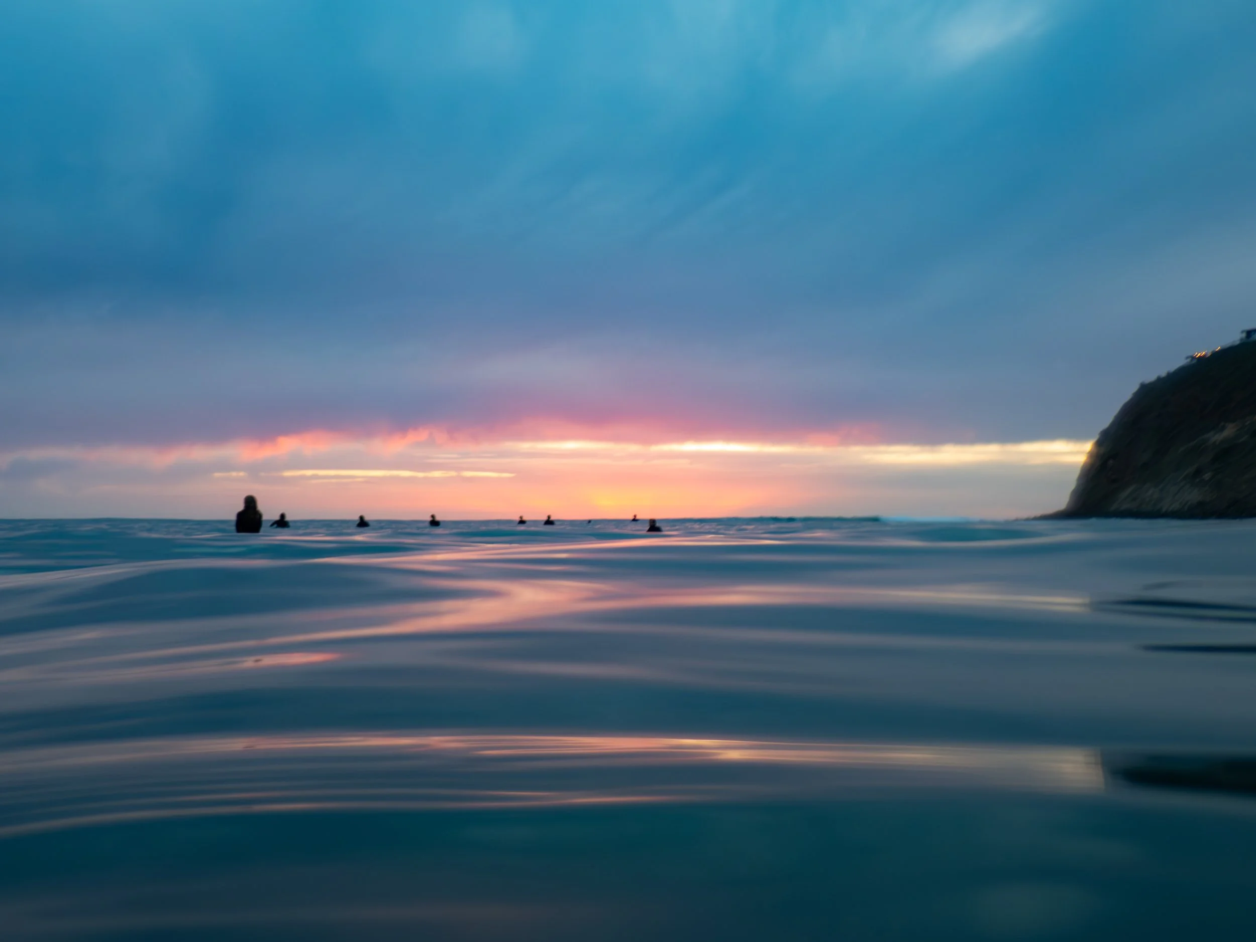 People surfing in the ocean during sunset with a cloudy sky and a rocky cliff on the right.