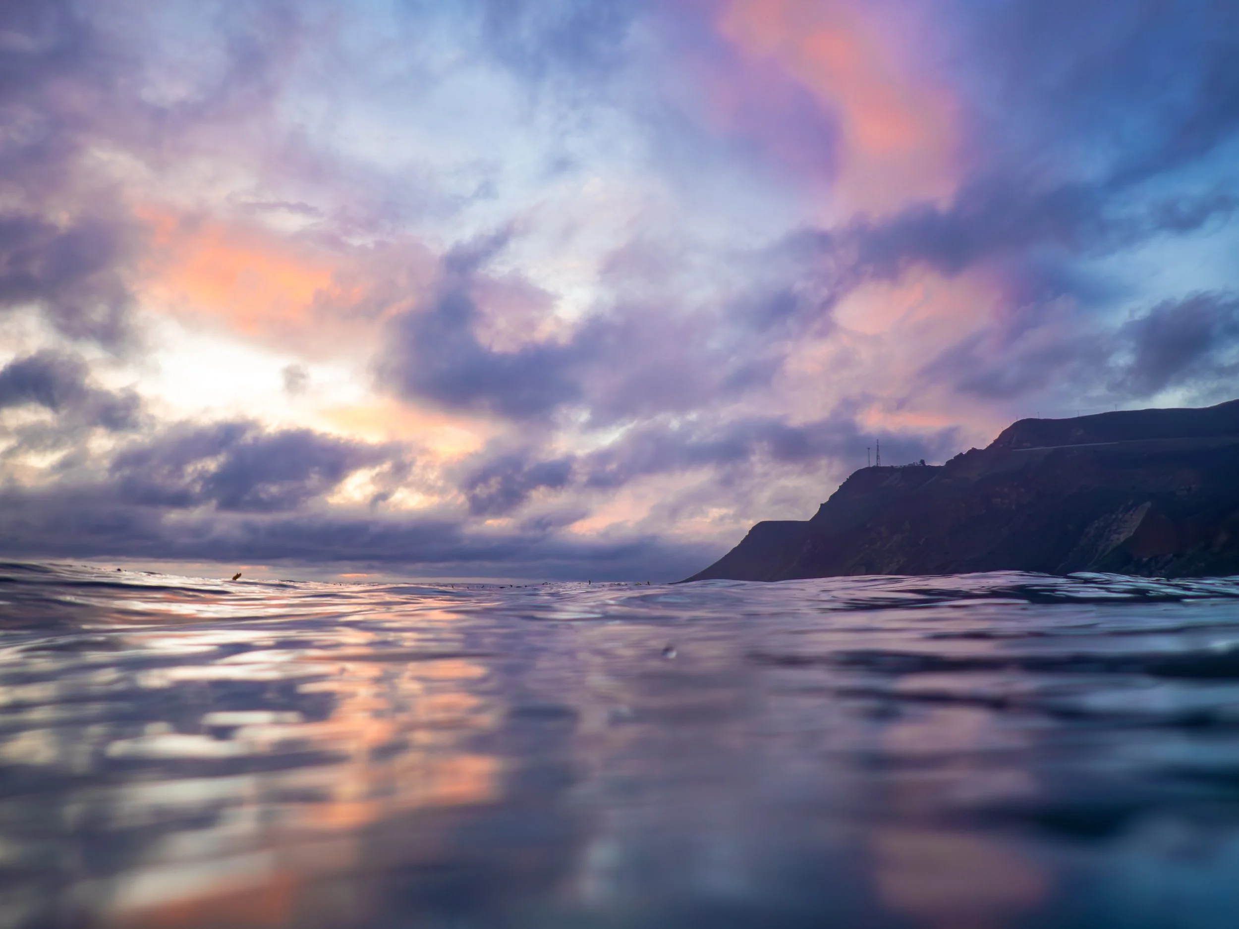 View of a cloudy sky during sunset above a calm sea with a silhouette of a mountain on the horizon.