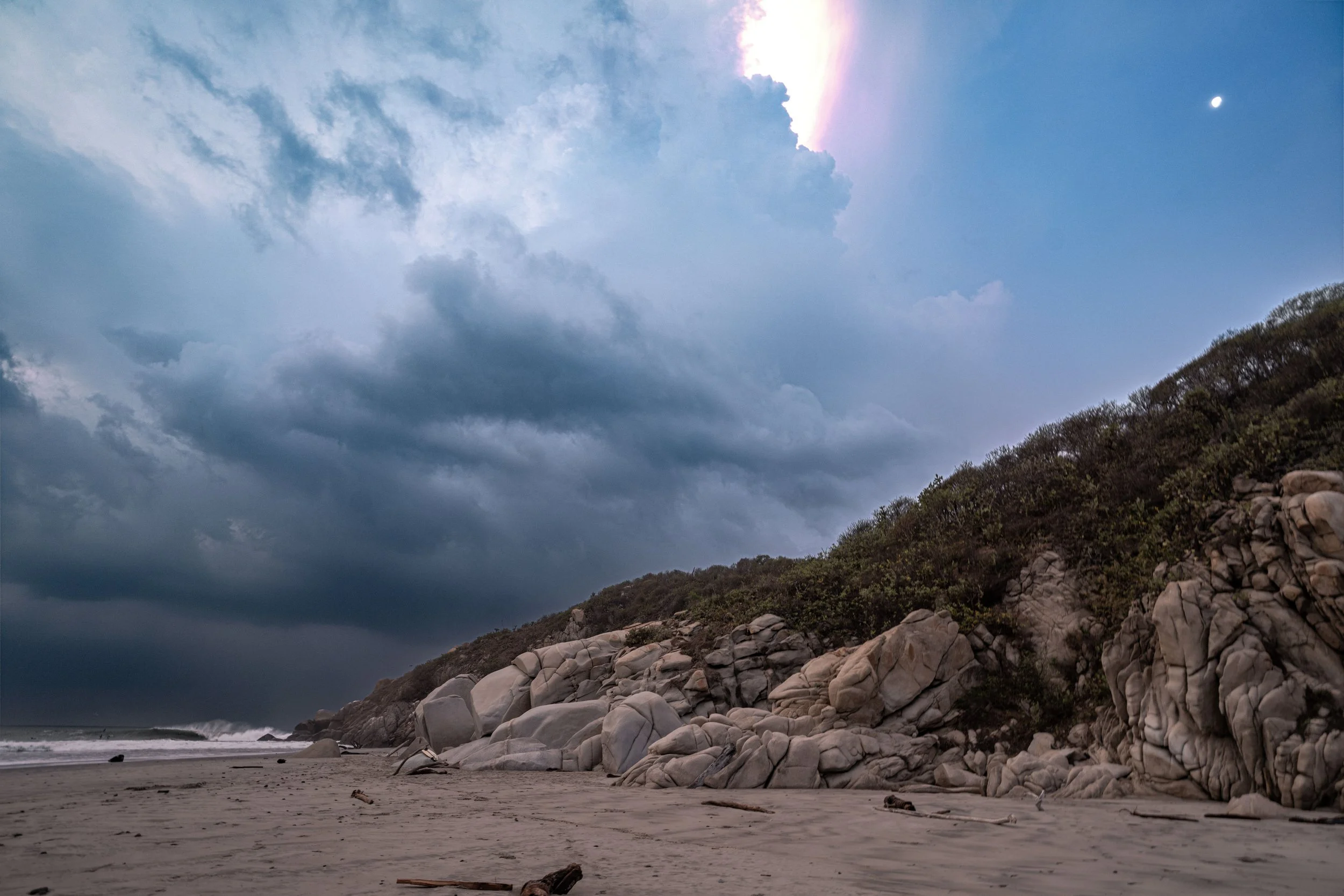 Coastal landscape with a cloudy sky, rocky shoreline, and hillside covered with bushes and rocks.