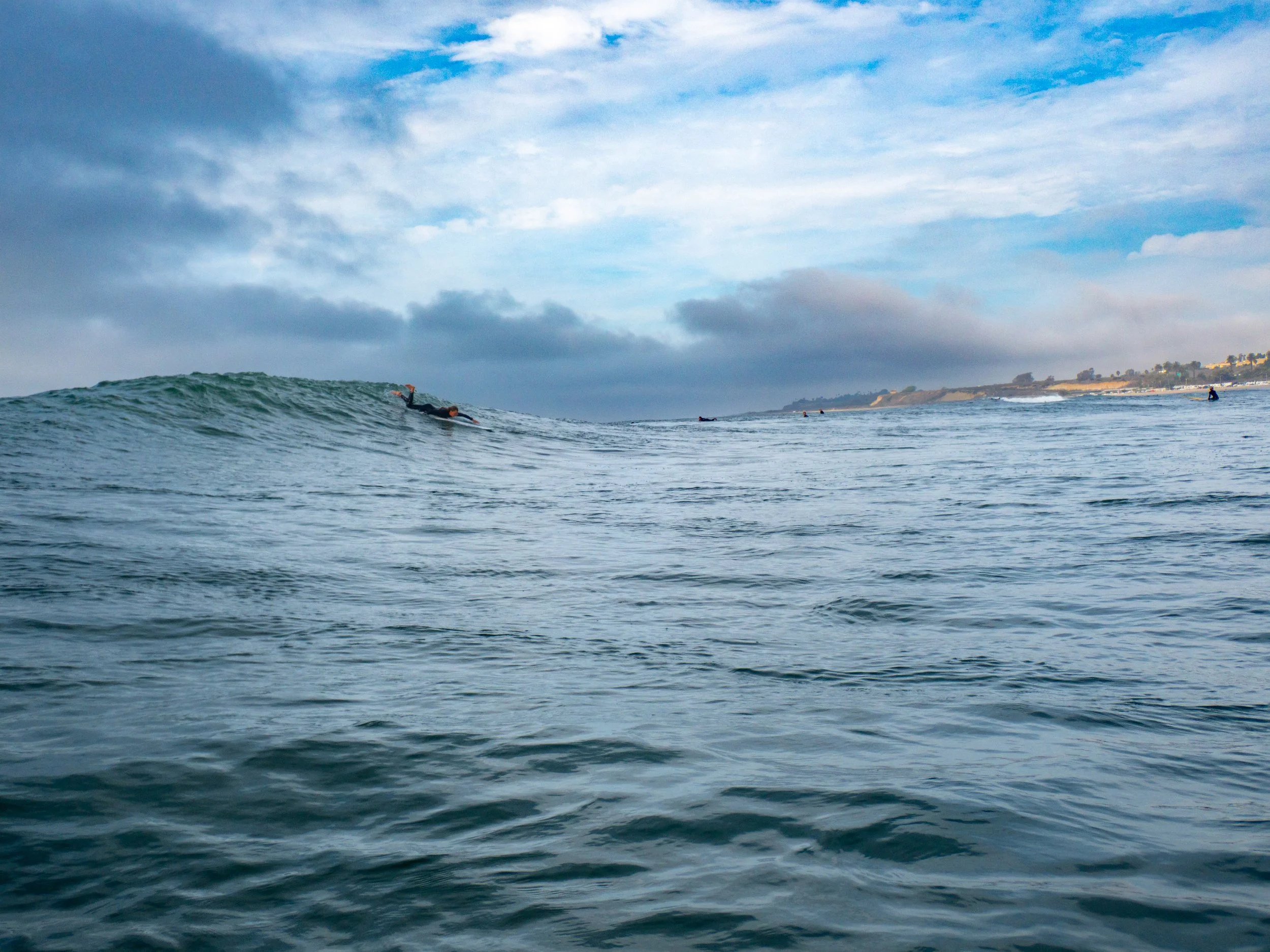 Surfer riding a wave in the ocean with a cloudy sky and distant shoreline in the background.
