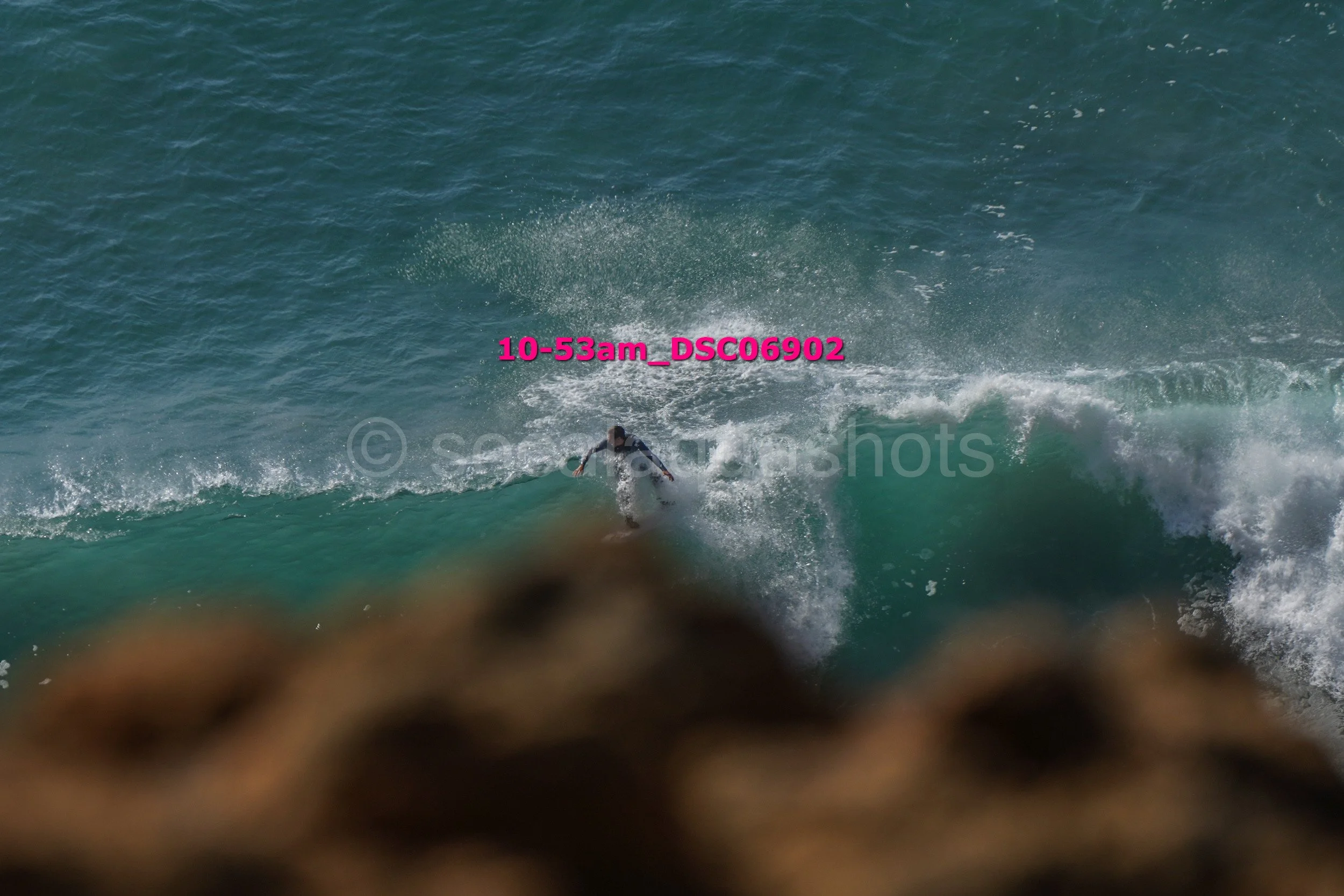 A person surfing on a blue wave in the ocean, with rocks in the foreground and some pink text overlayed on the image.