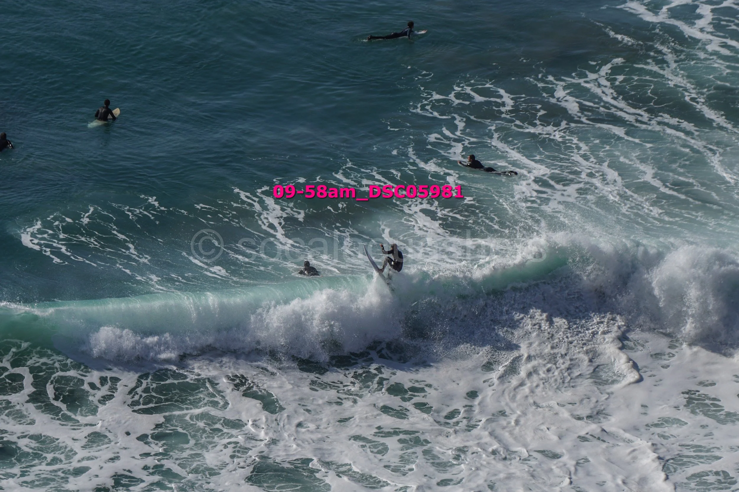Surfer performing a trick on a wave with several surfers waiting in the water.