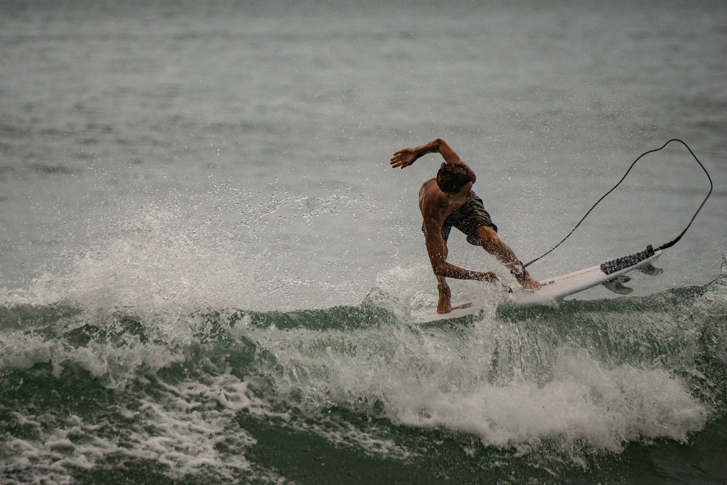 A person surfing on a wave in the ocean, leaning forward on a surfboard, with water spray around, and an overcast sky.