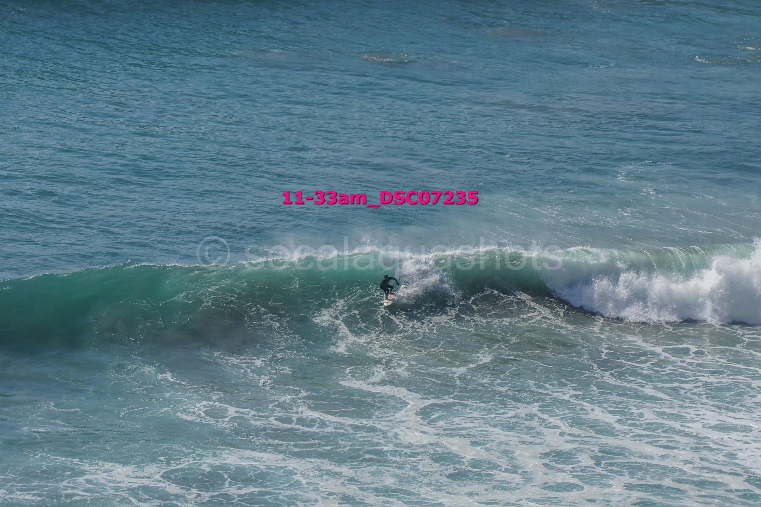 A person surfing on an ocean wave with blue water and white foam, wearing a wetsuit.