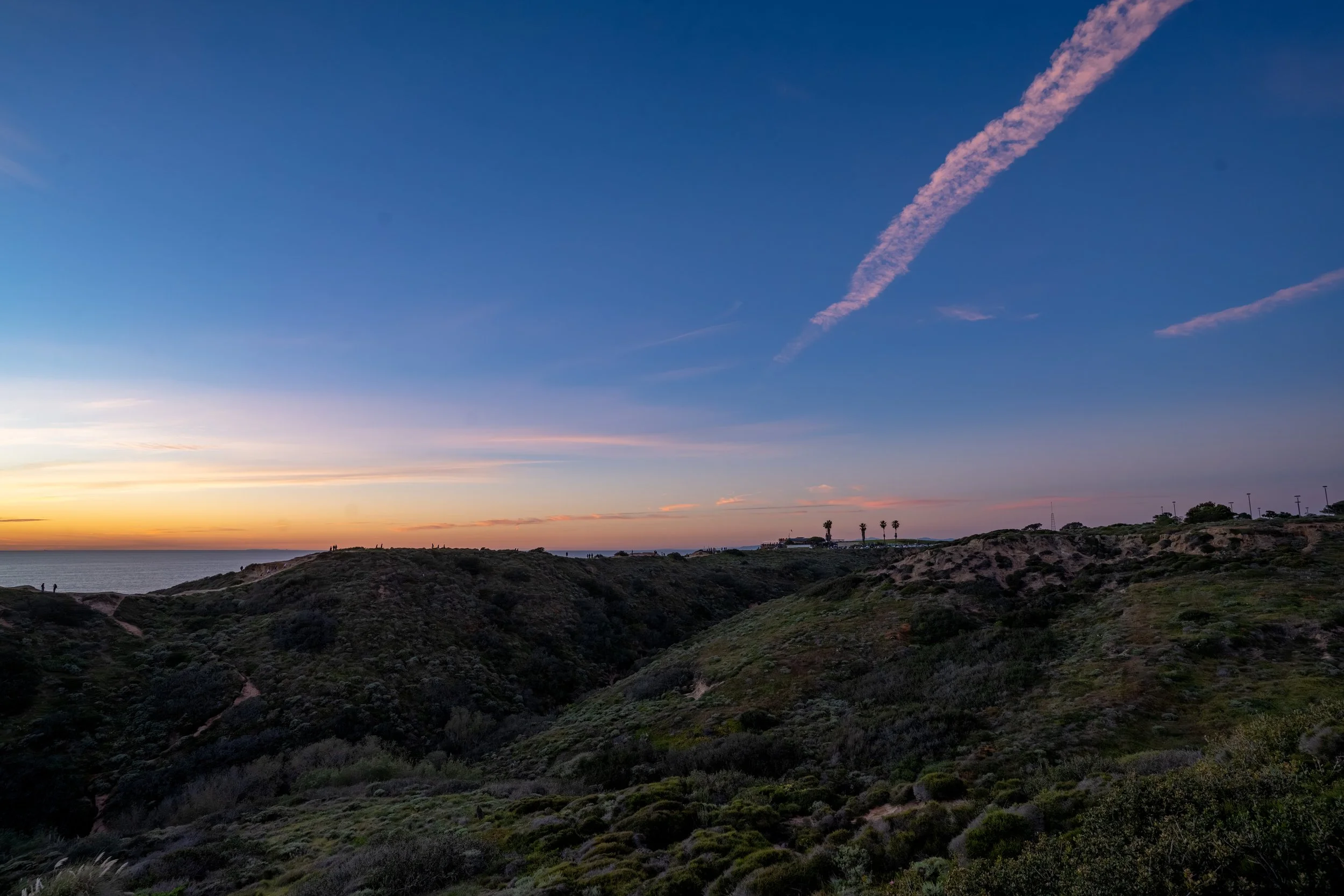 Coastal hills at sunset with a partly cloudy sky and contrails.
