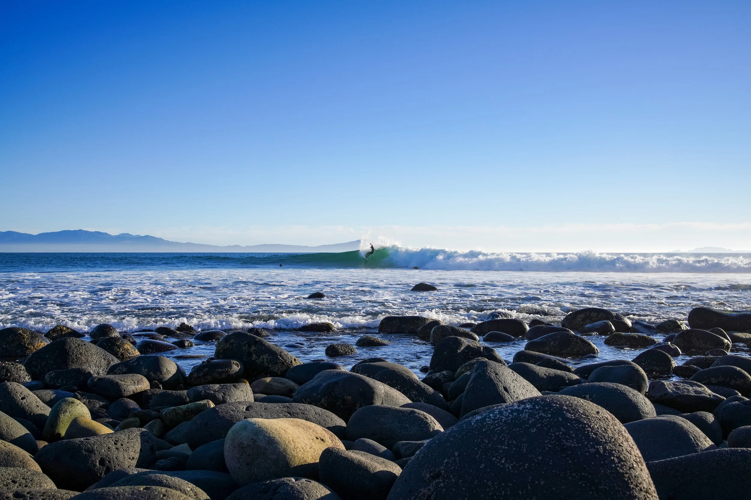 A person surfing on a wave at a rocky beach with mountains in the background under a clear blue sky.
