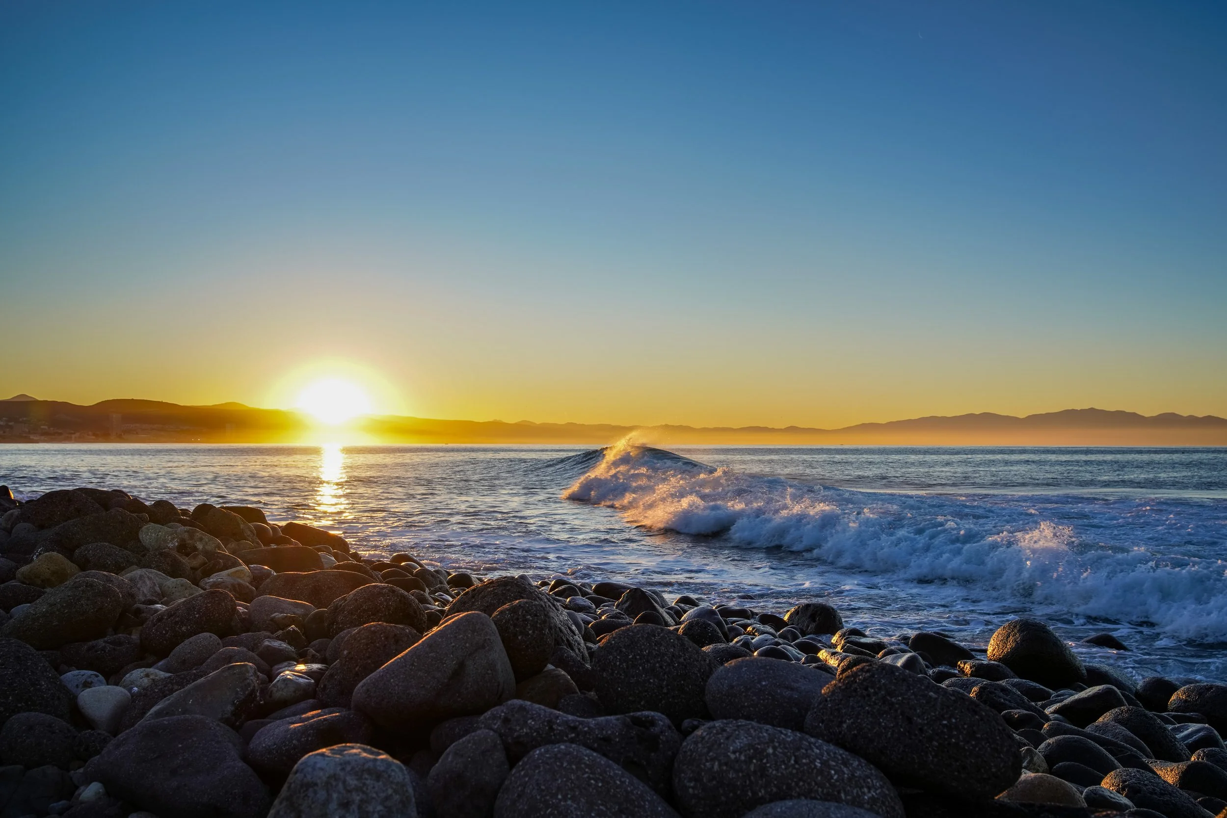 Sunset over the ocean with waves crashing onto a rocky shore, mountains in the background, and a clear sky.