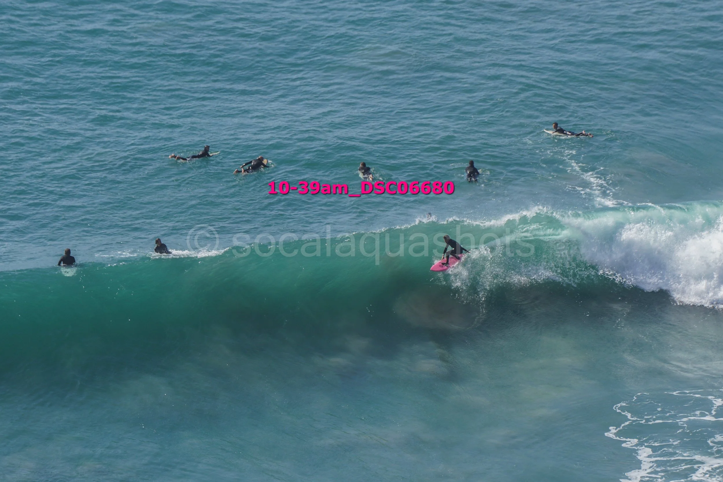 A group of people surfing in the ocean, with one person riding a pink surfboard on a wave and others paddling or swimming nearby.