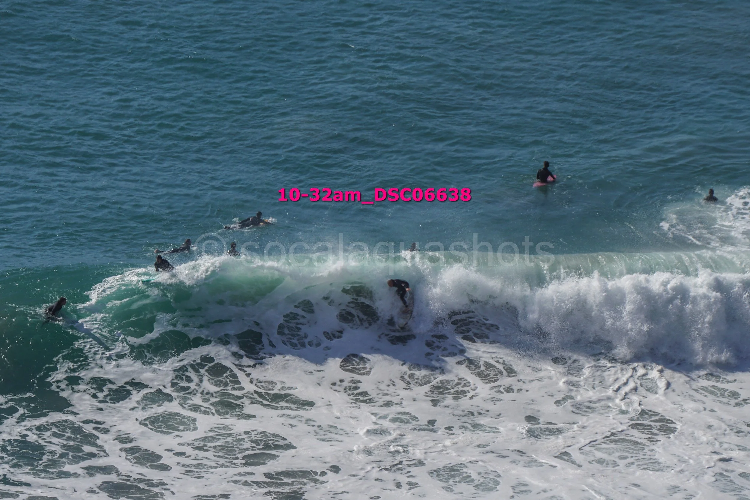 Surfers in black wetsuits riding and waiting for waves in the ocean, with one surfer near the crest of a wave.
