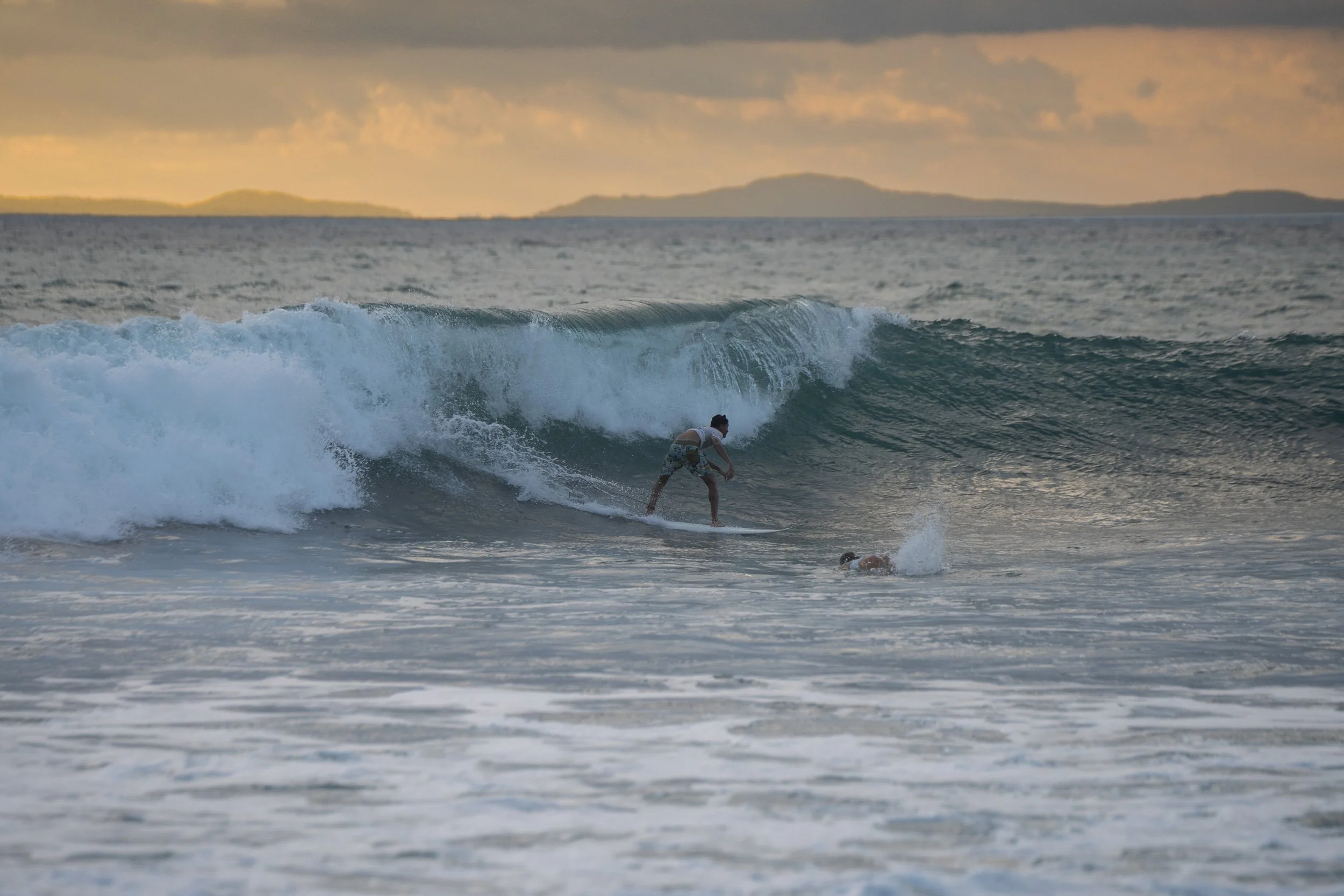A person surfing on a wave in the ocean during sunset or sunrise, with islands or land visible in the background and dark clouds above.