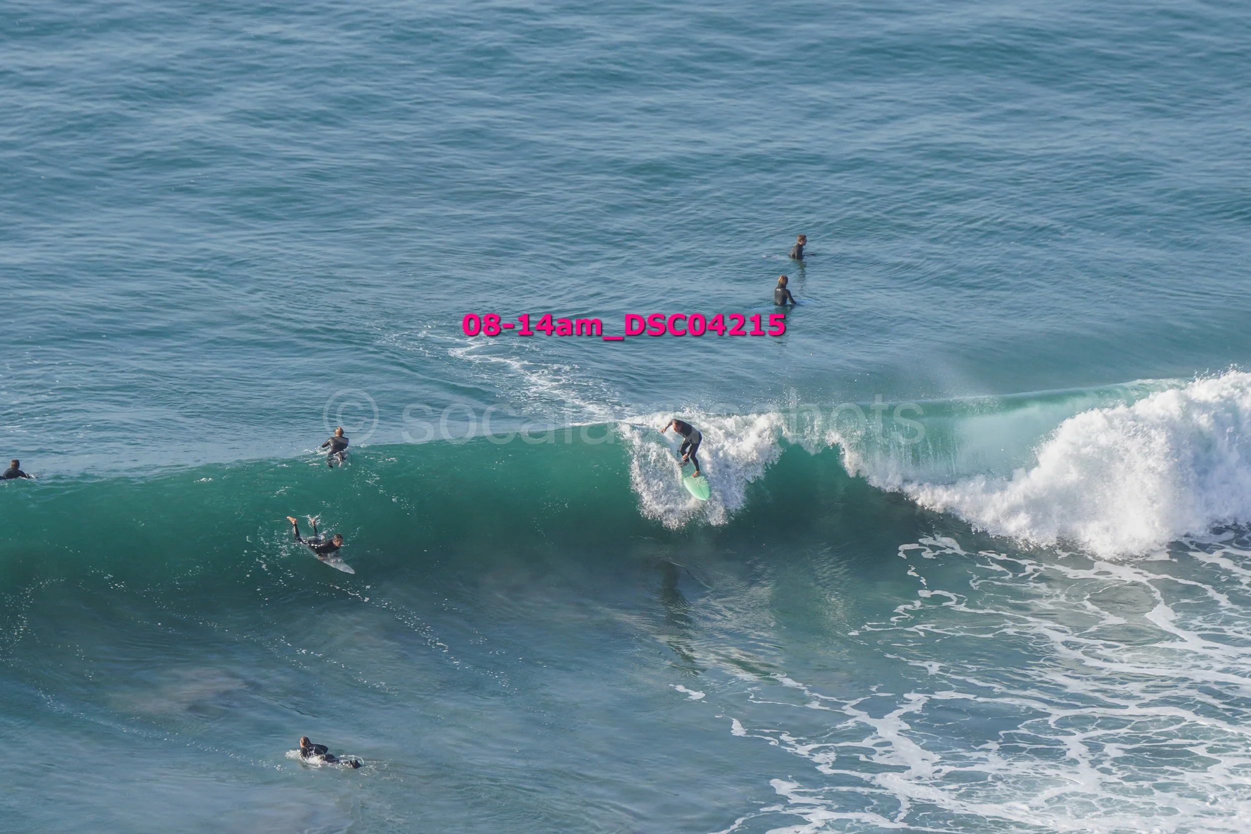 Surfer riding a wave with several surfers in the water nearby on a sunny day.